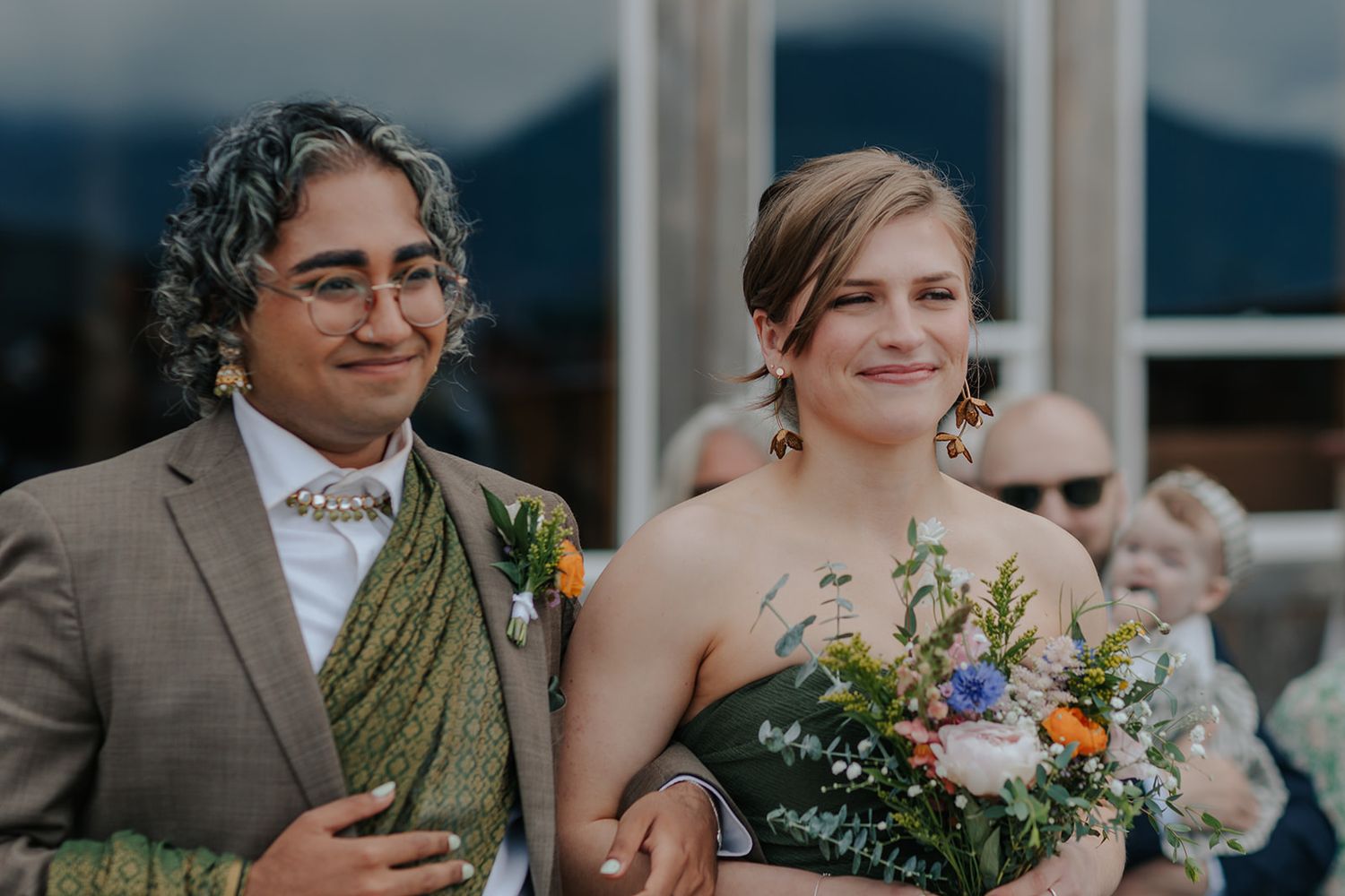 Wedding couple poses for photos in formal attire with colorful bouquet against mountain backdrop.