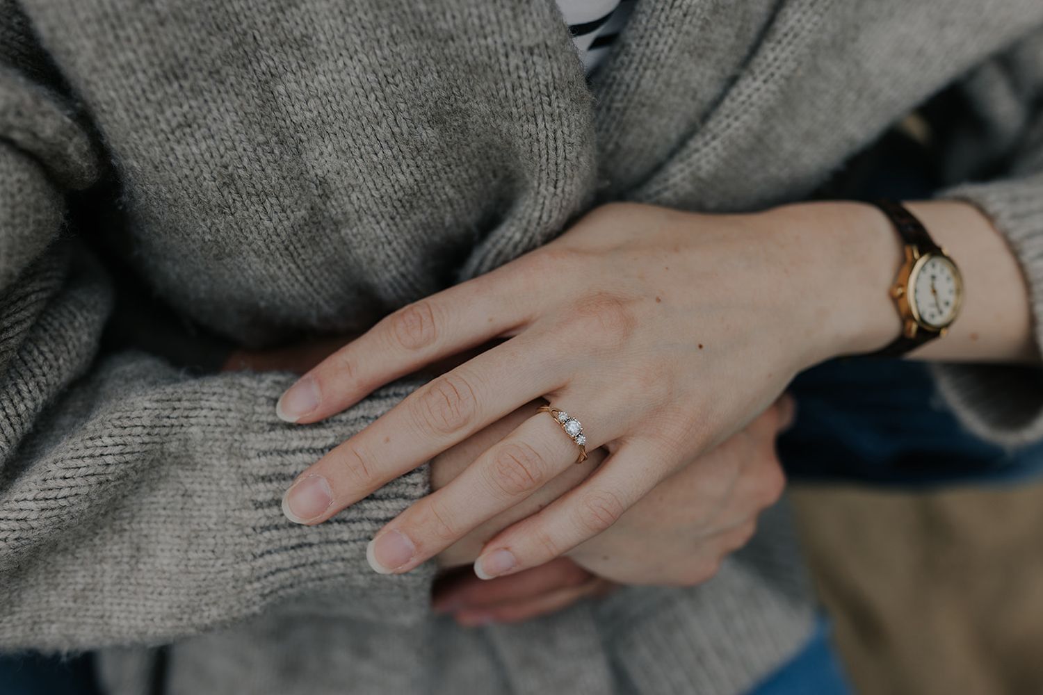 Hand wearing an engagement ring resting on a gray sweater in natural light.