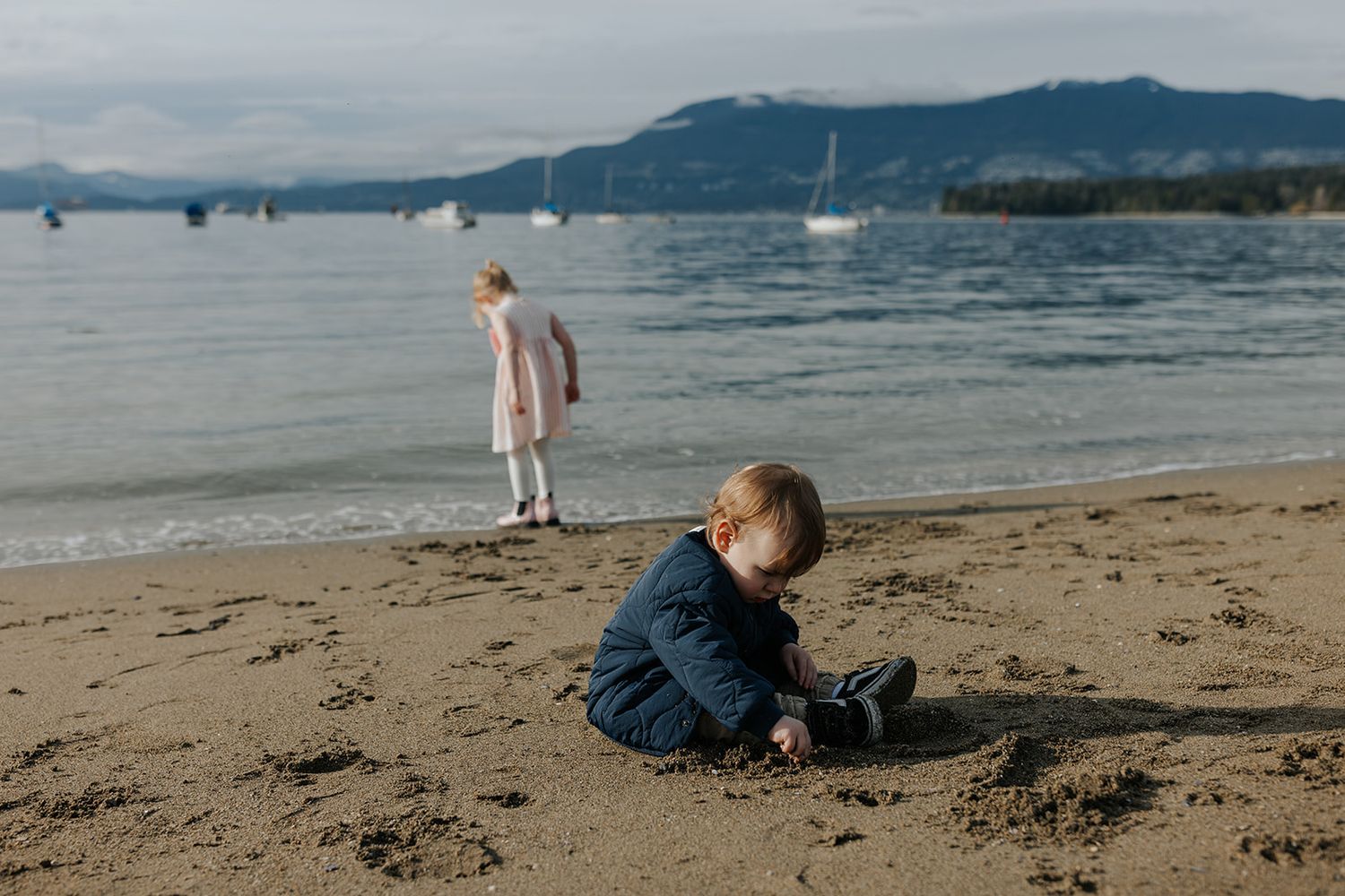 Children playing on a sandy beach with boats and mountains visible in the distance.