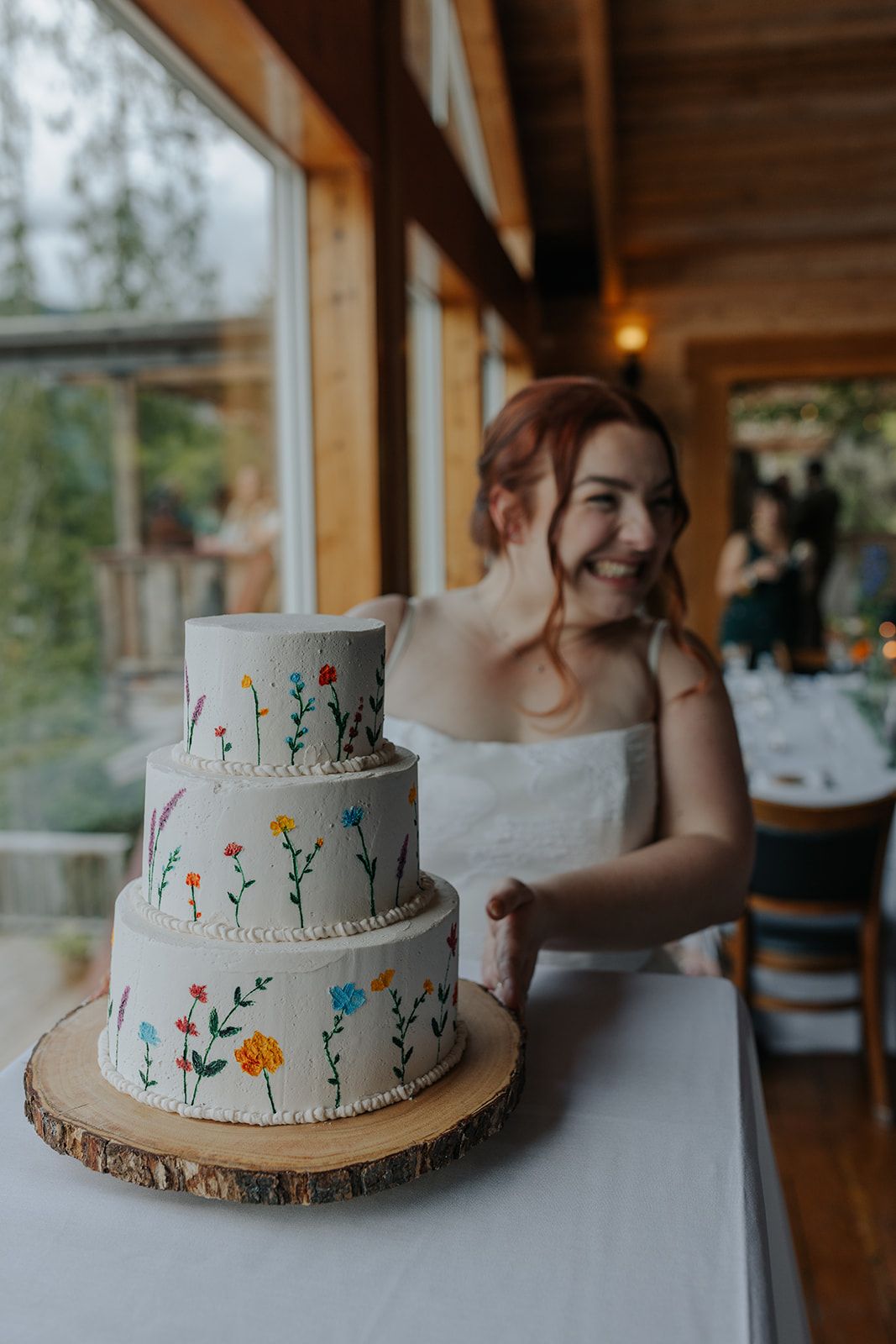 Three-tiered wedding cake decorated with colorful hand-painted wildflowers on wooden stand.