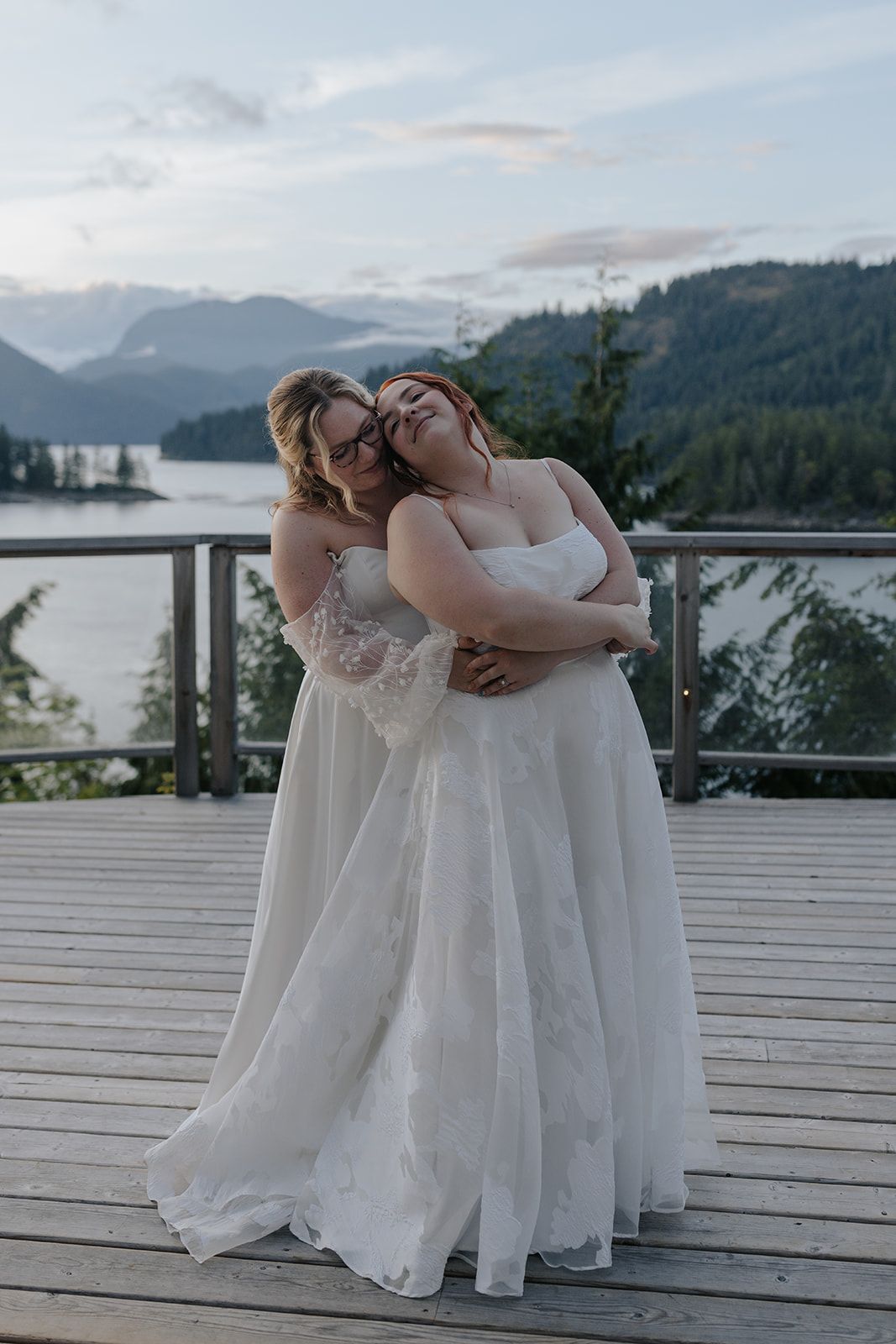 Wedding party members in white dresses huddle together on a deck with mountain scenery.