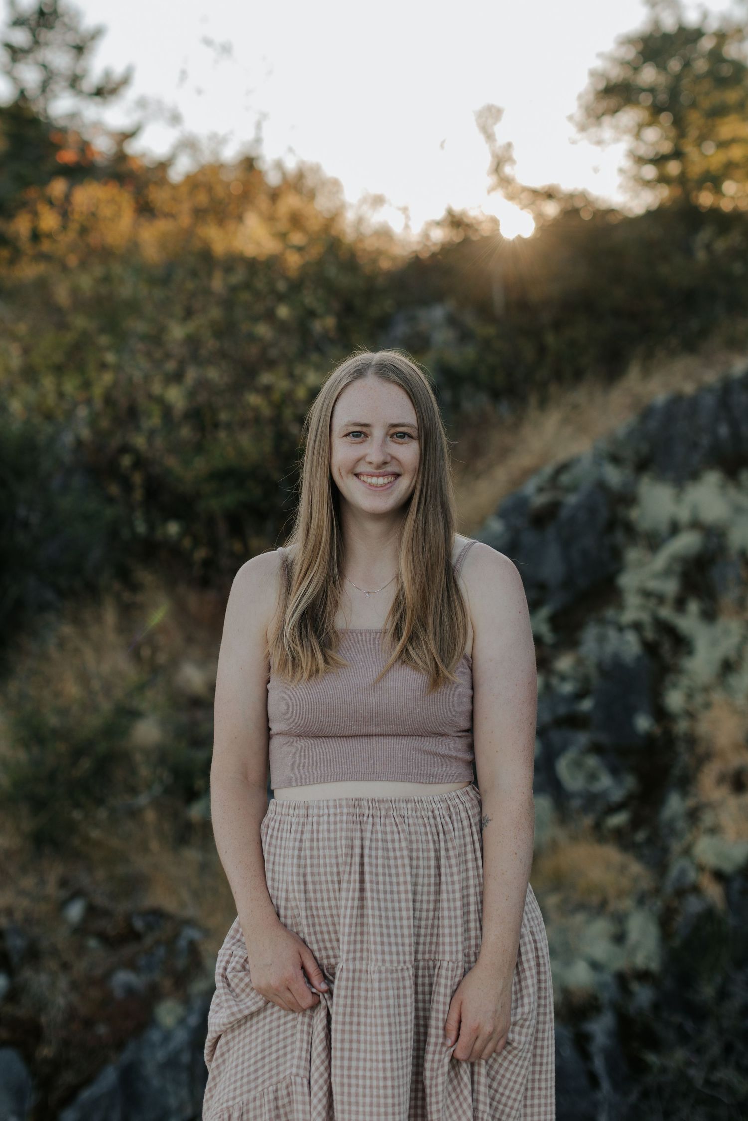 Woman in checkered dress poses outdoors during sunset with natural scenery.