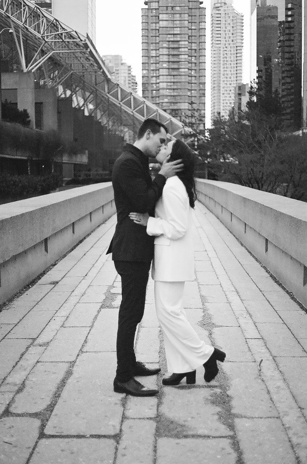 A romantic couple embraces on a bridge with city buildings in the background.