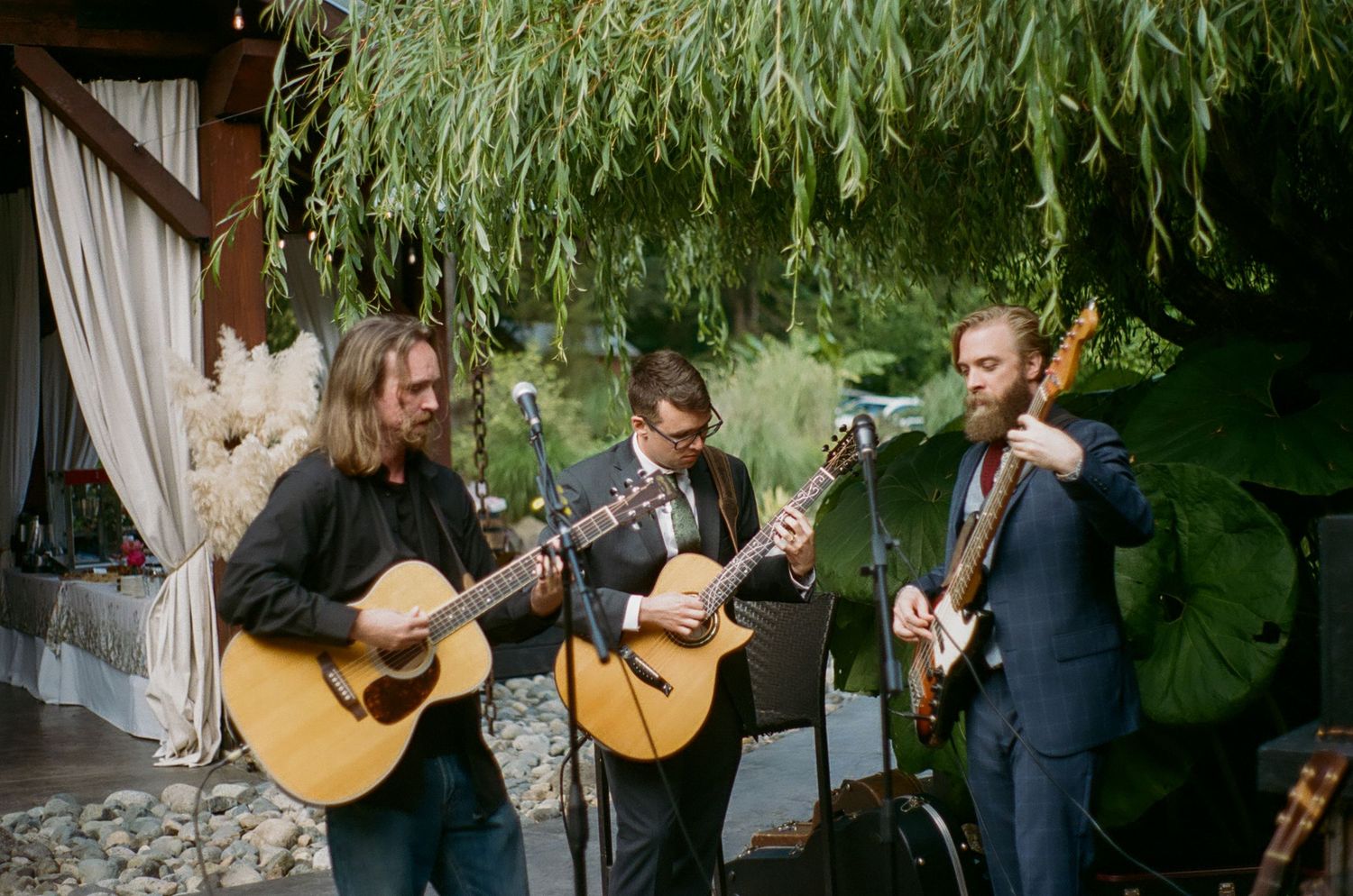 Musicians perform with acoustic guitars under a covered outdoor venue with hanging greenery.