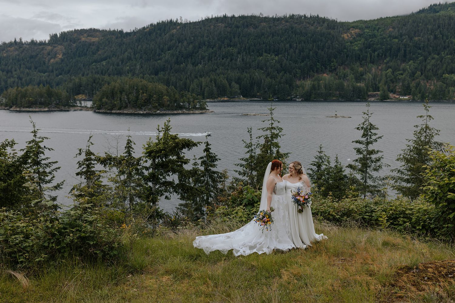 Two wedding figures embrace on a hilltop overlooking a scenic mountain lake landscape.