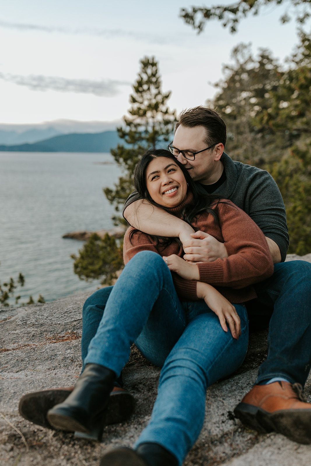 A couple shares a tender moment while sitting on rocky shoreline overlooking a lake.