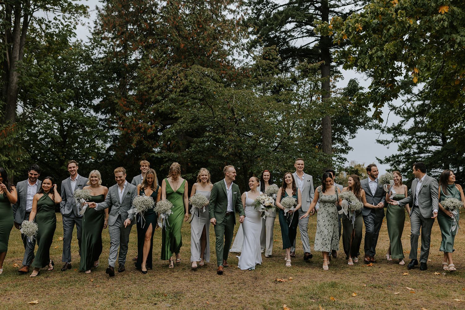 Wedding party poses for a group photo on grassy hillside with trees in background.