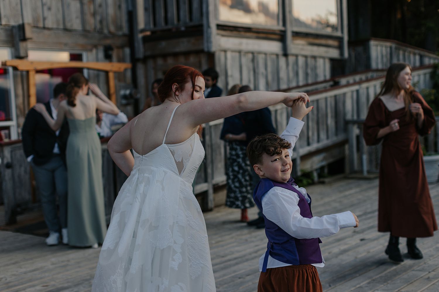 A person in a white wedding dress dances with a young child in traditional dress on a wooden deck.