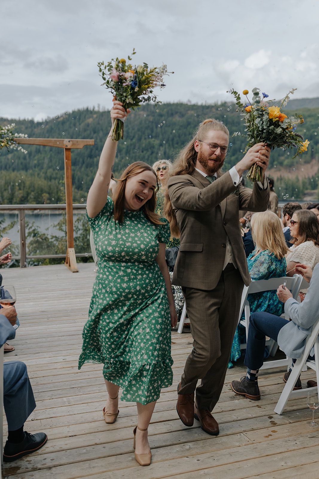 Couple in green dress and brown suit dancing playfully on wooden deck with mountain backdrop.