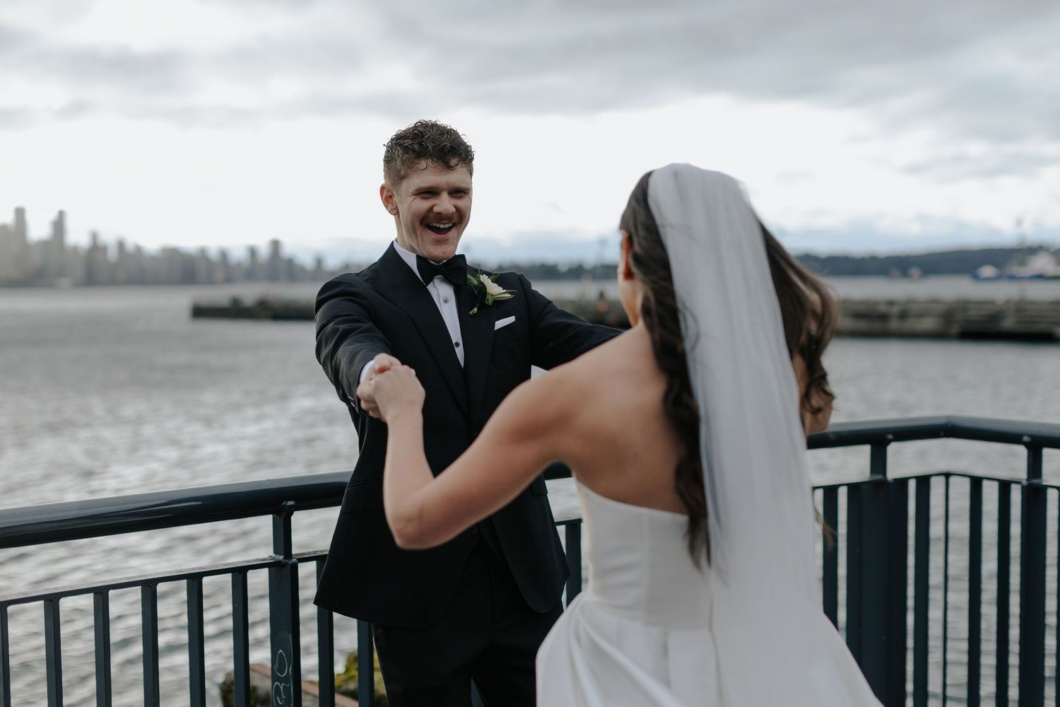 A couple shares a joyful moment during their wedding celebration on a waterfront deck overlooking a city skyline.
