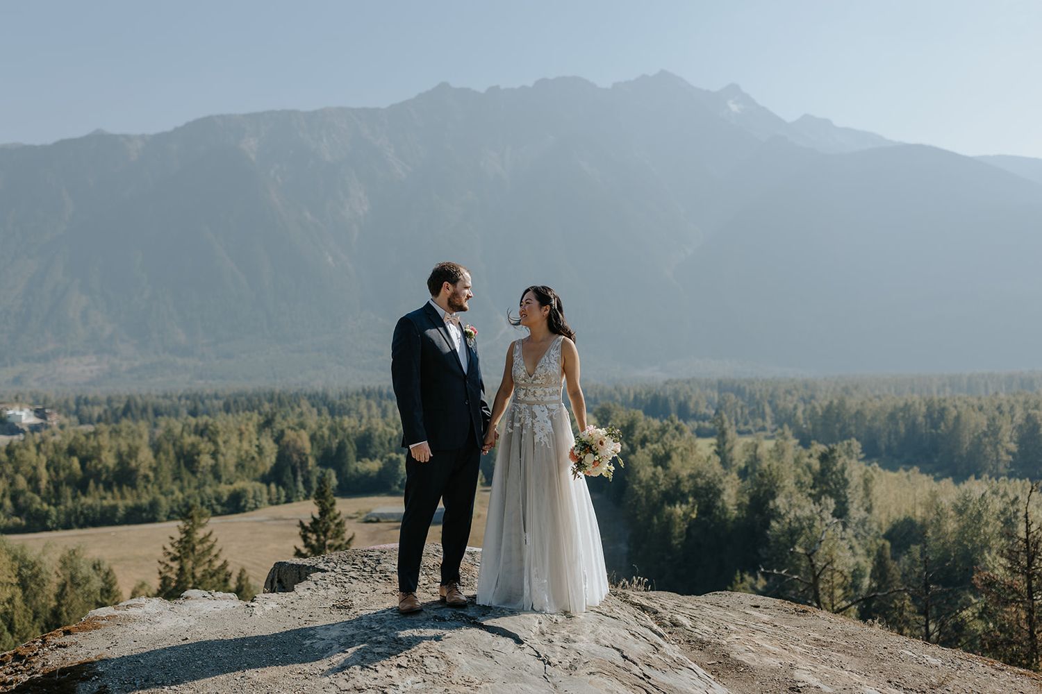 A couple stands on a rocky mountain overlook with evergreen forests and majestic peaks in the background.