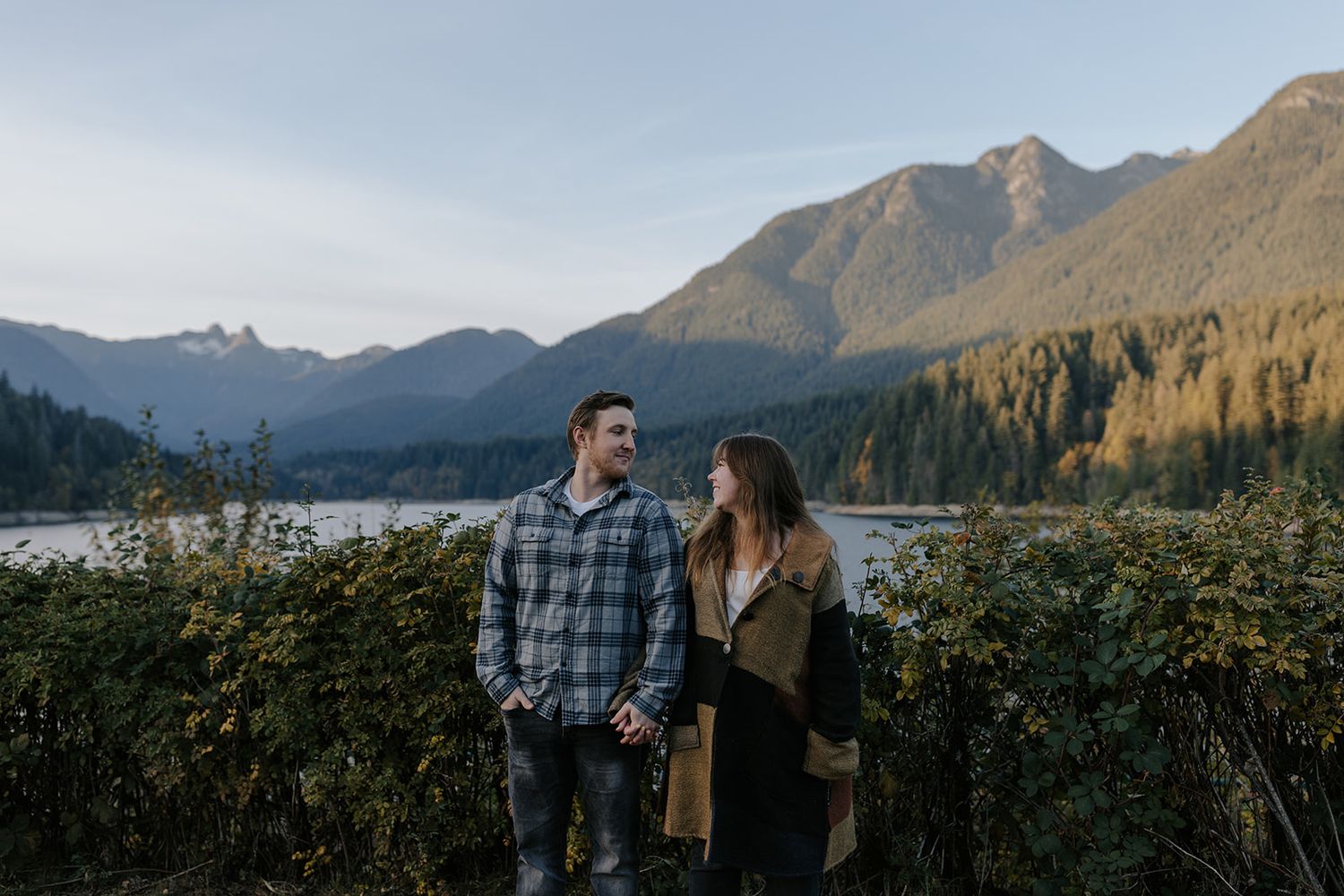 Couple standing together by a scenic mountain lake surrounded by lush greenery and forested peaks at golden hour.