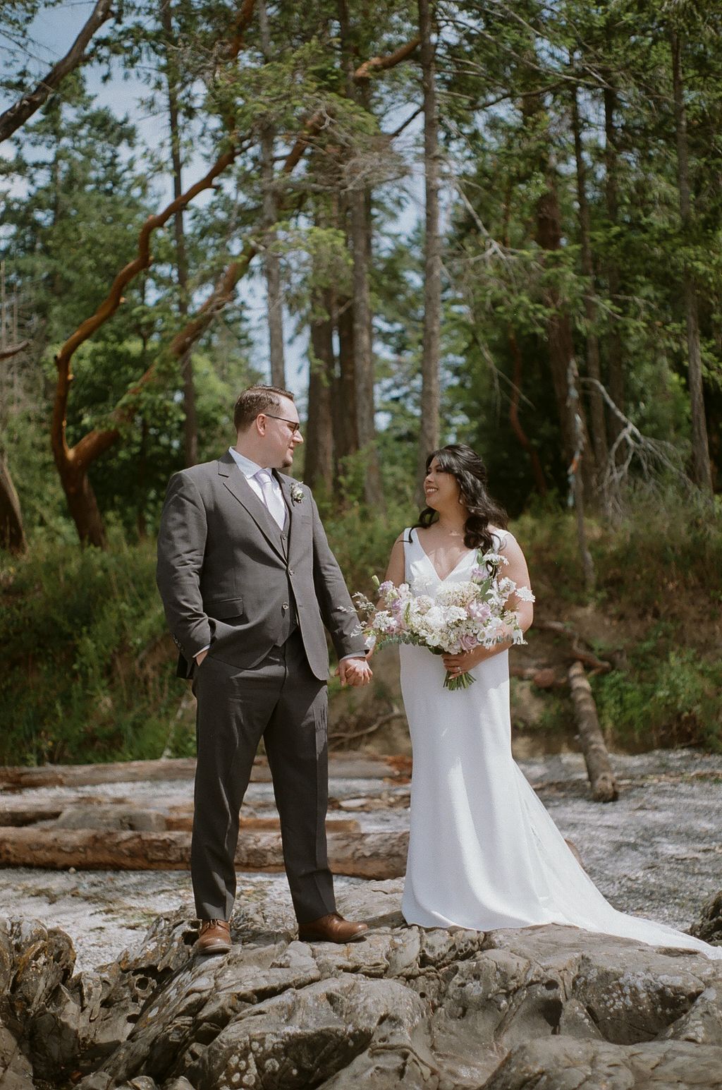 A wedding couple stands on rocky terrain surrounded by tall evergreen trees.