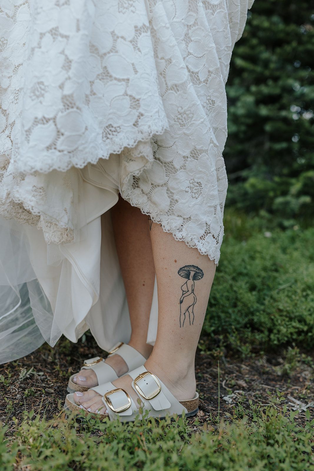 Close-up of bride's feet in white sandals with delicate lace dress hem and small tattoo visible.