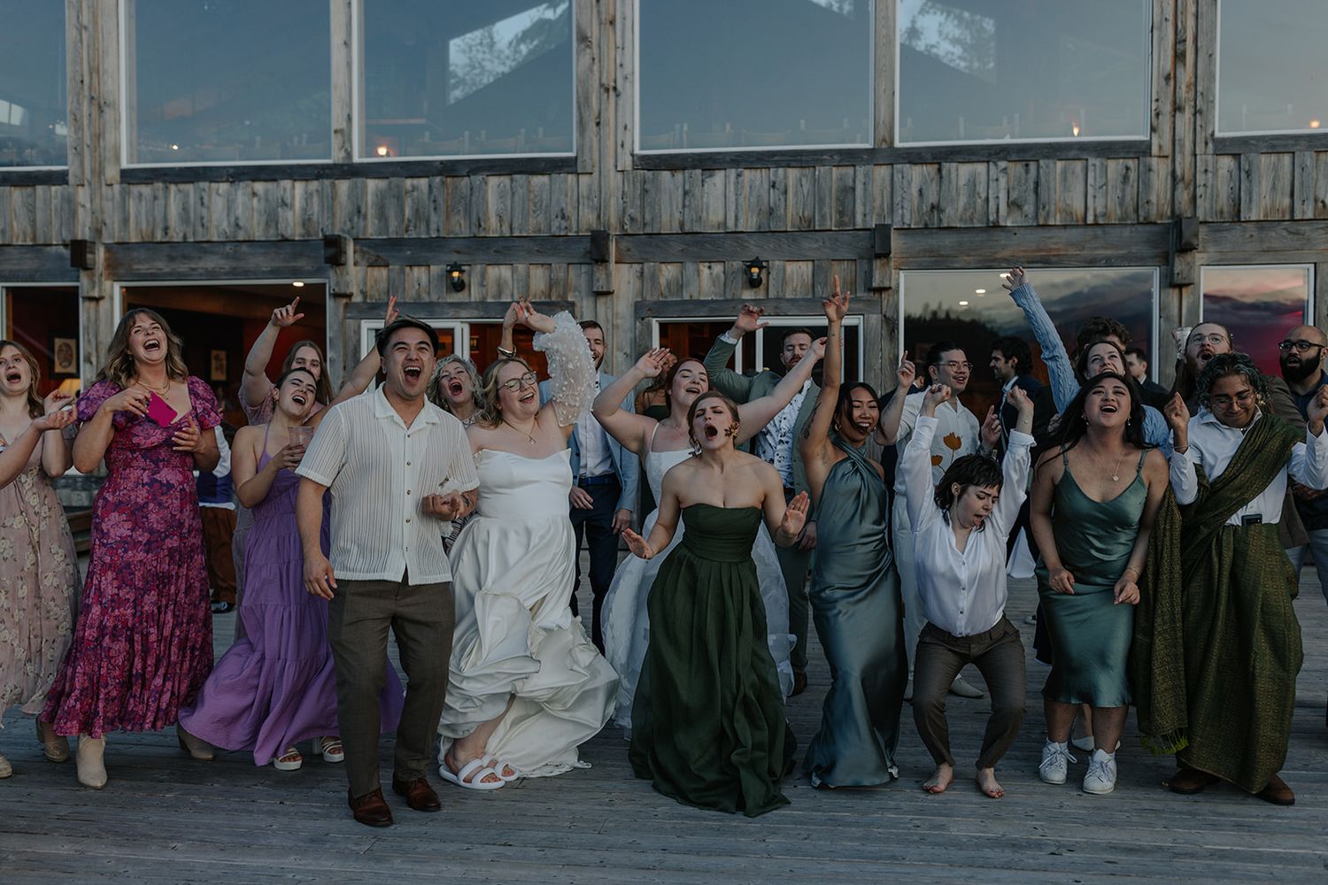 Wedding party celebrates joyfully on a pier with raised arms and colorful attire.