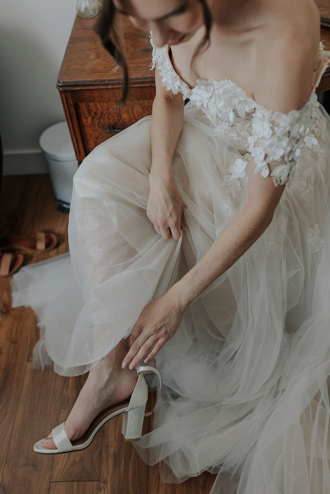 Close-up details of a bride putting on white wedding shoes with delicate tulle dress fabric.