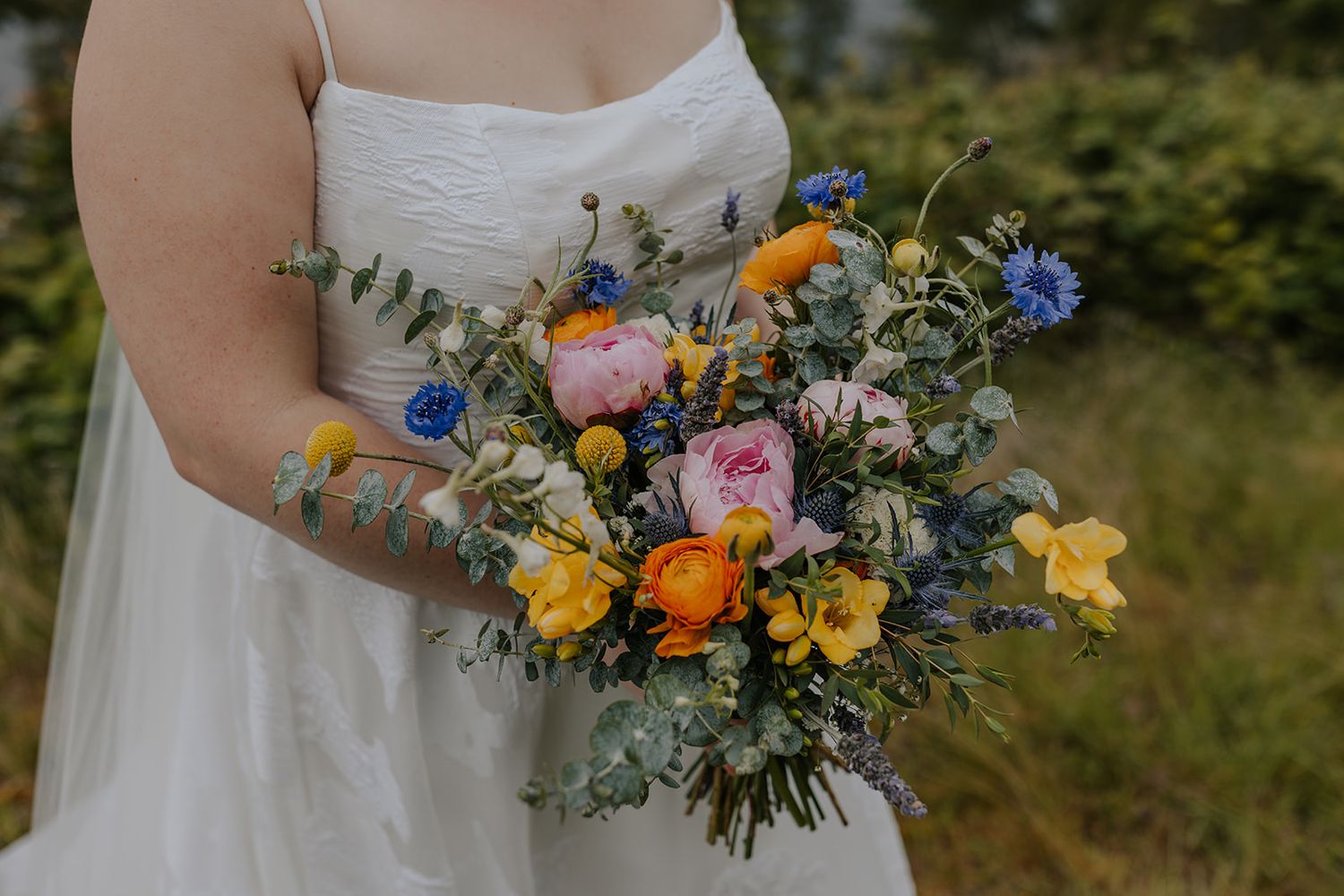 Close-up of a wildflower wedding bouquet with blue, orange and pink blooms held against a white dress.