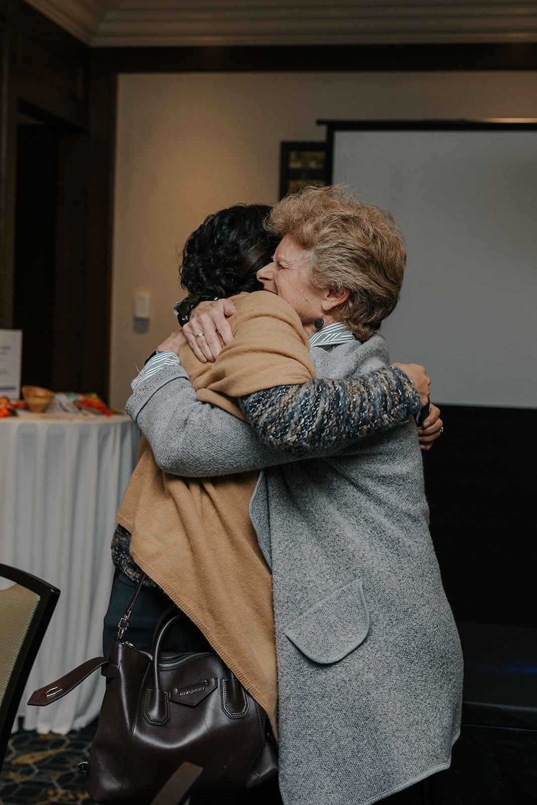 People embracing in an indoor setting wearing grey and tan coats.