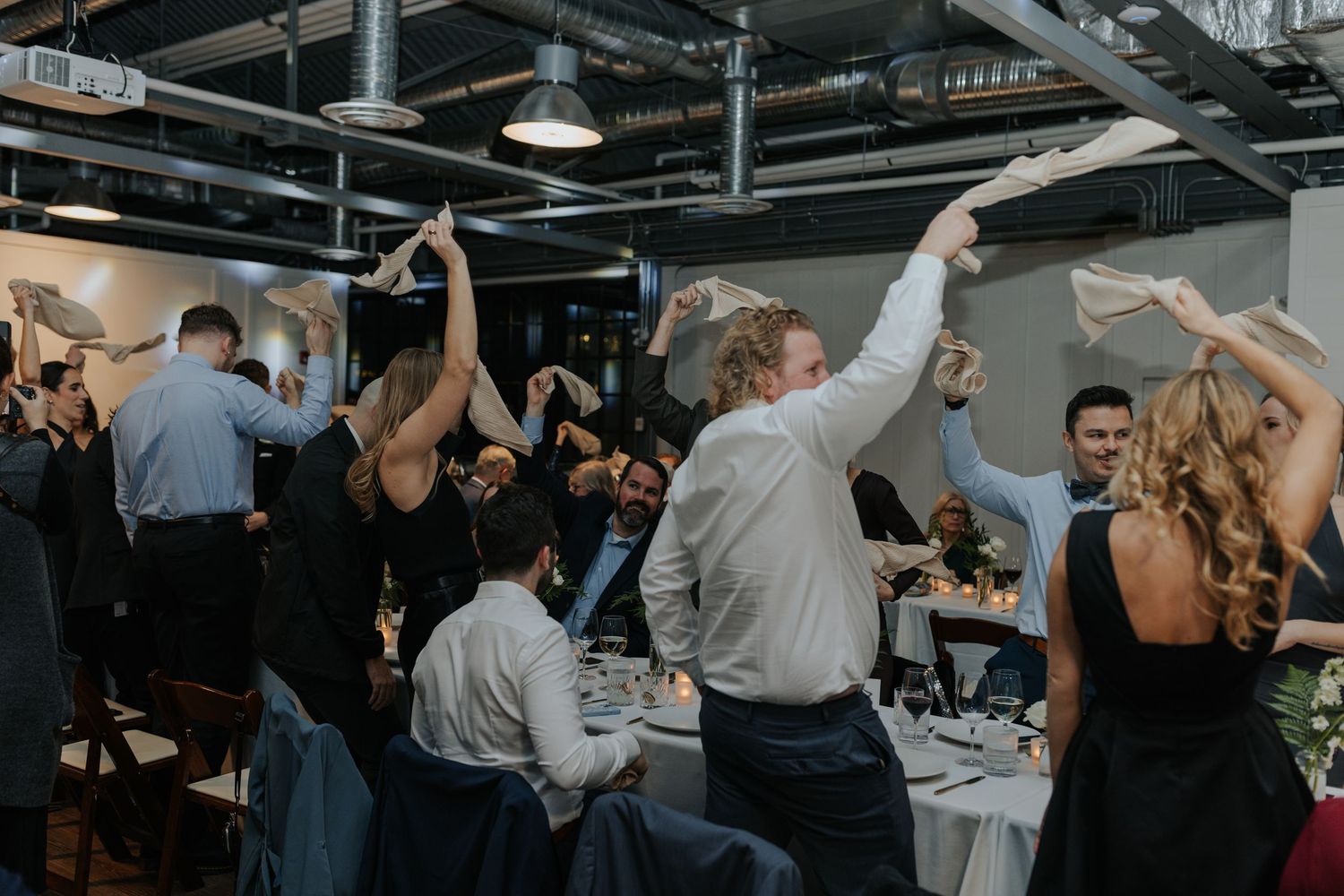 Wedding reception guests dancing and celebrating on a wooden dance floor under dim lighting.