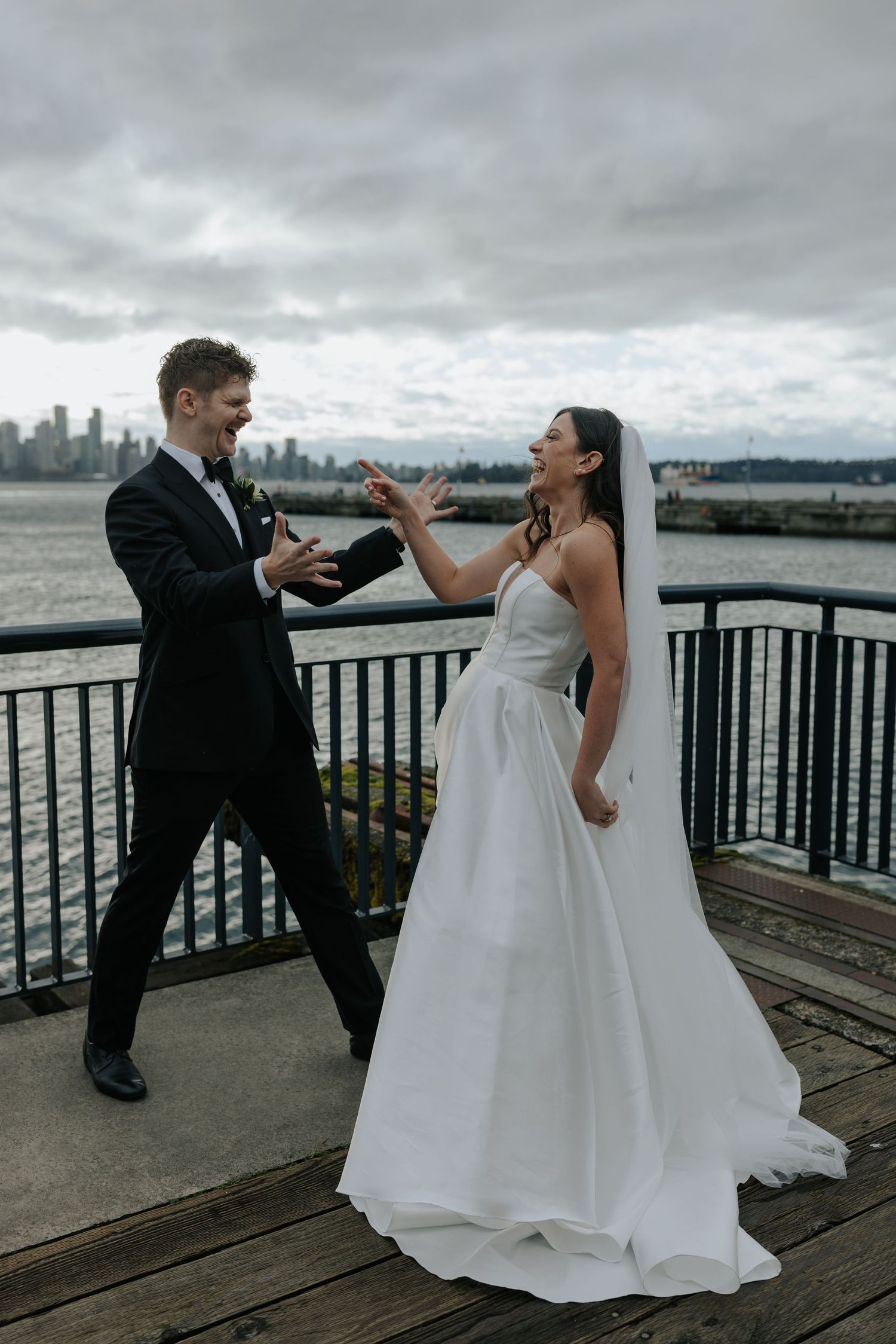 A couple in wedding attire playfully interact on a waterfront deck with a cloudy cityscape backdrop.