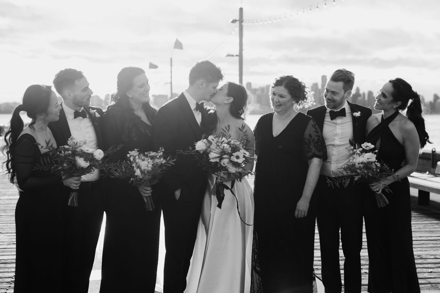Wedding party members stand in a row with arms linked on a dock by the water at sunset.