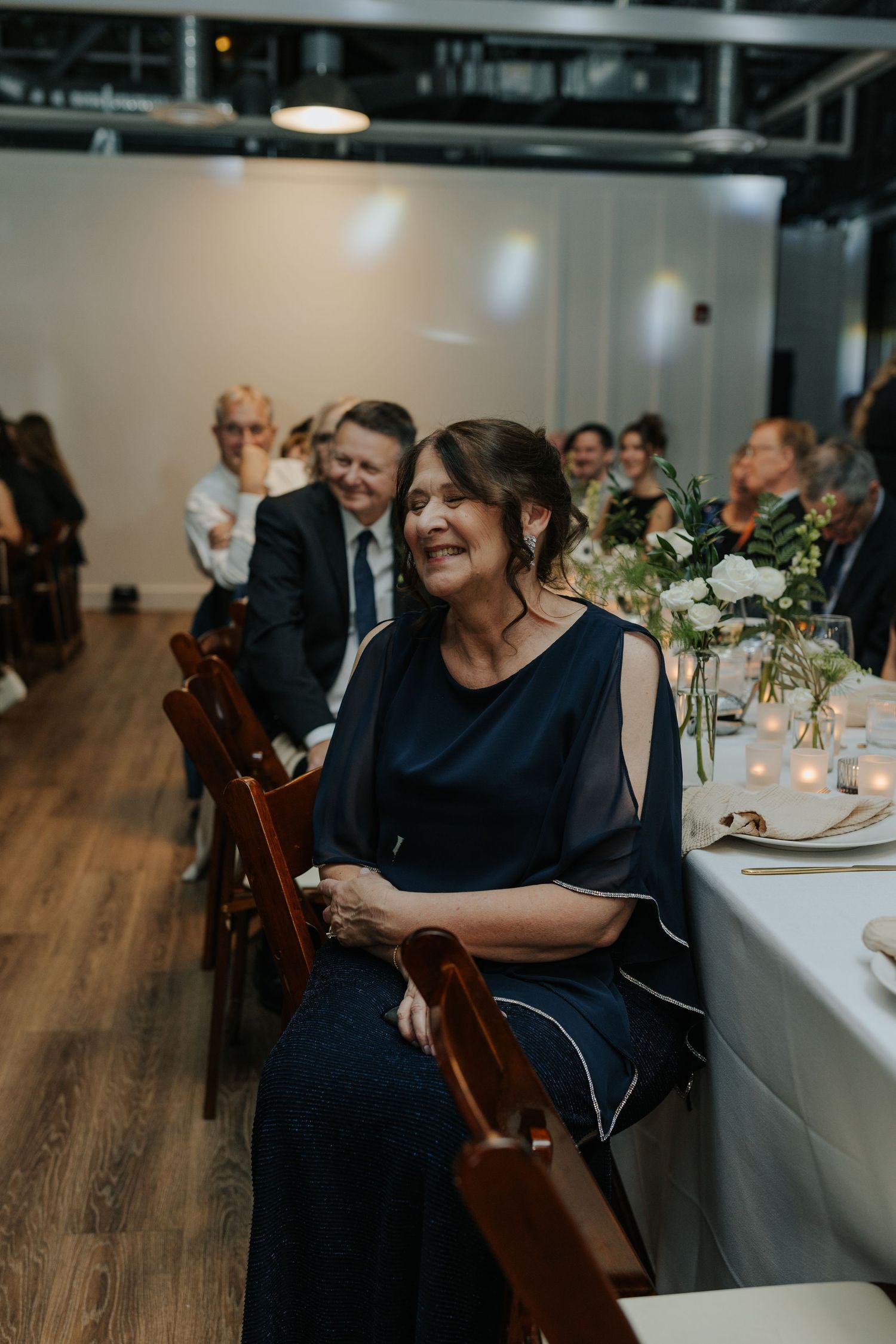 Wedding guest in navy blue dress smiles while seated at reception with elegant table settings and hardwood floors.