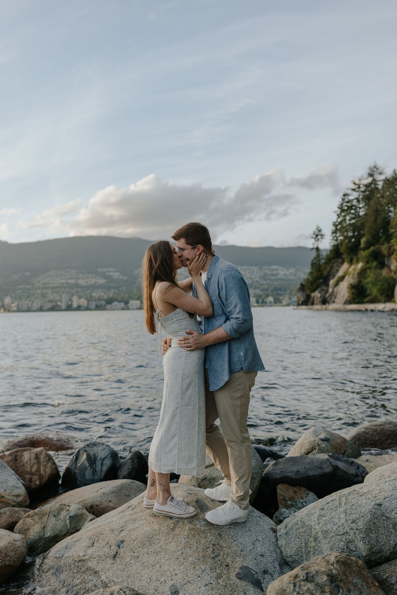 Couple kissing on rocky shoreline with mountain lake backdrop, woman in gray dress, man in blue blazer.