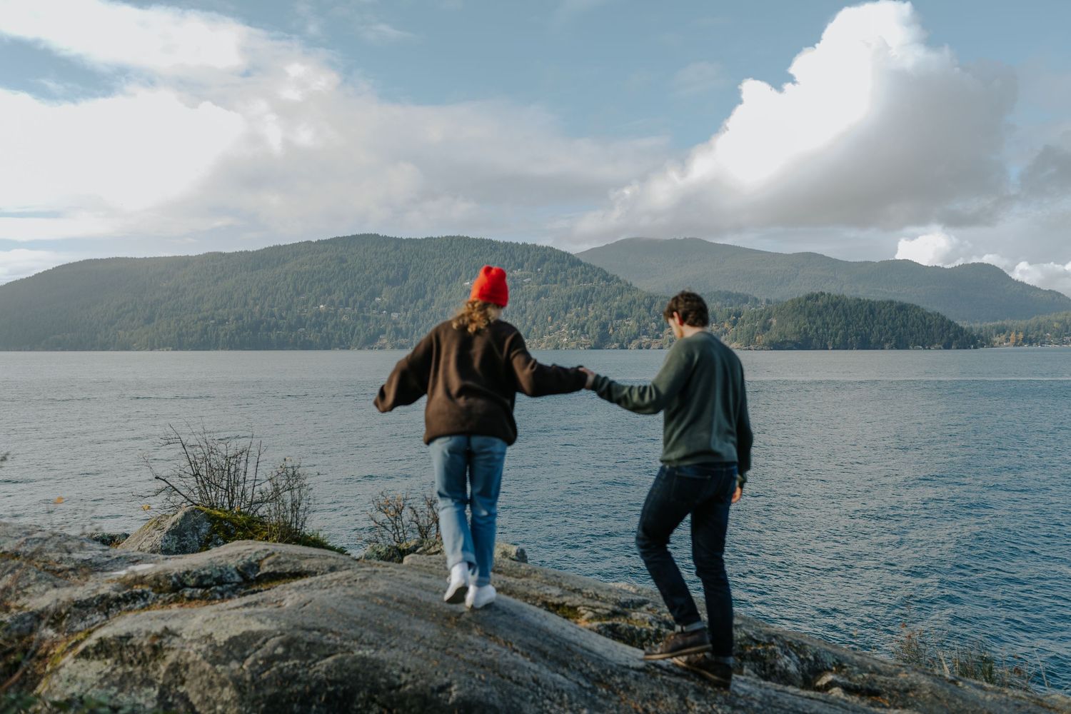 Couple holding hands while standing on rocky shore overlooking scenic mountain lake under cloudy sky.