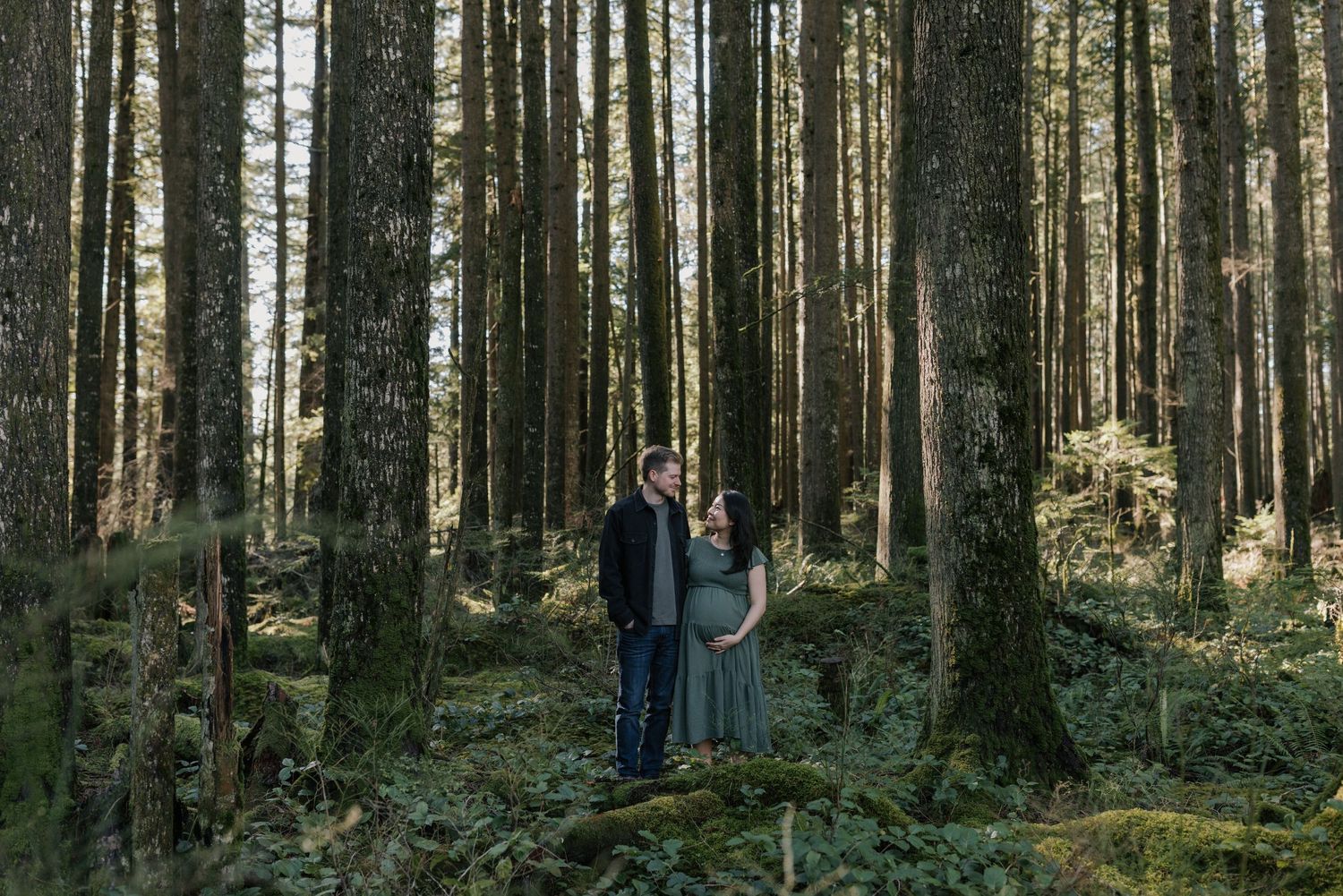 Couple shares a tender moment in a lush green forest, surrounded by tall trees, during a maternity photo session.
