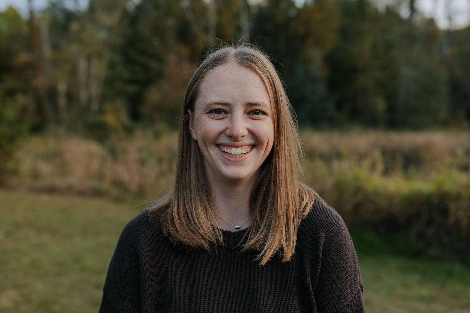 Smiling young woman with straight brown hair wearing a black top, standing outdoors in a natural field setting.