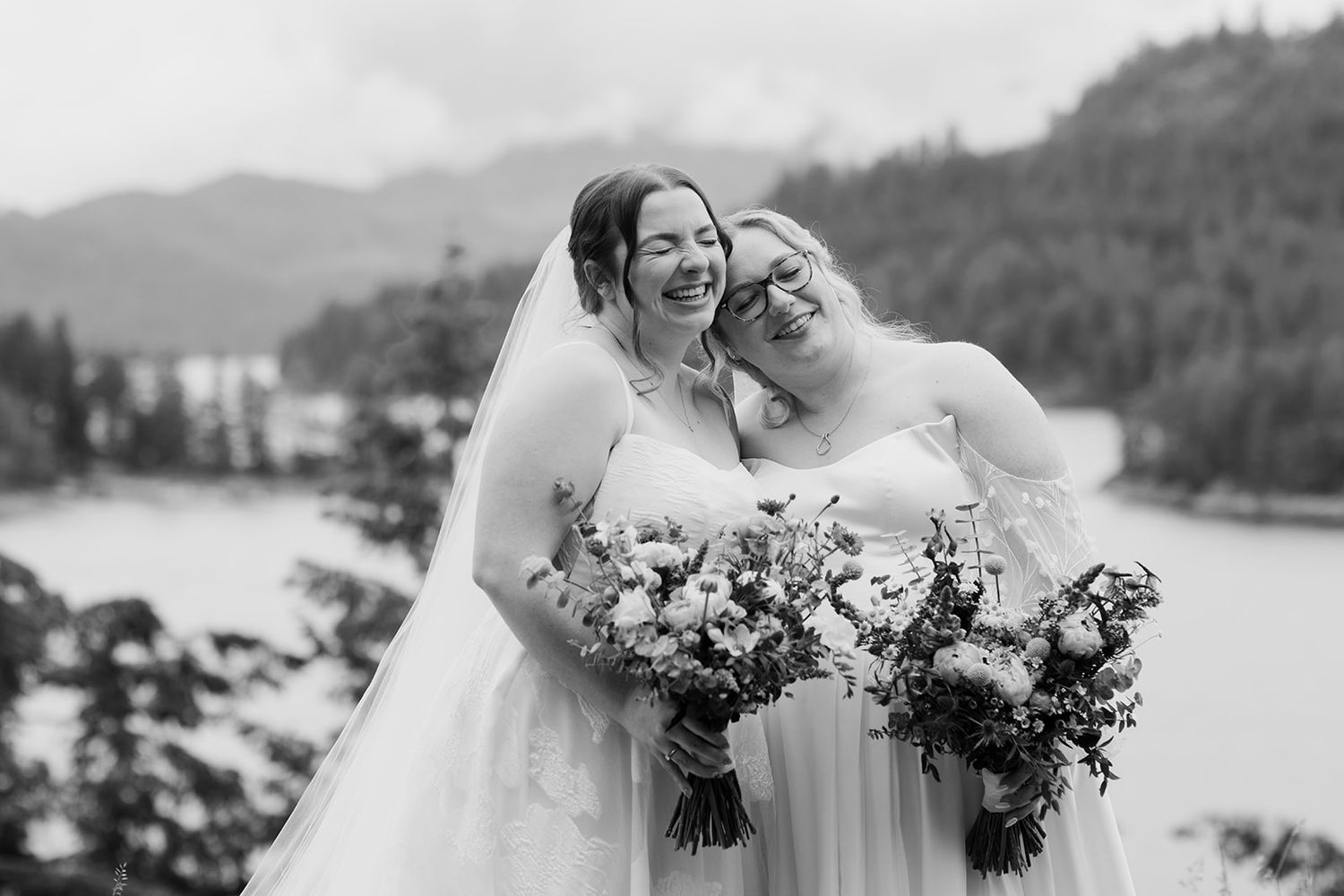 A black and white portrait of two people in wedding dresses sharing a laugh by a lake.