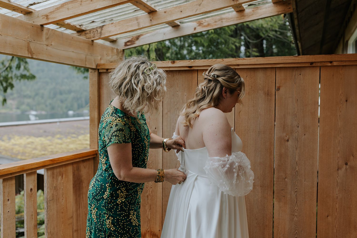 A person in an emerald green dress helps adjust a white wedding gown on a covered outdoor deck.
