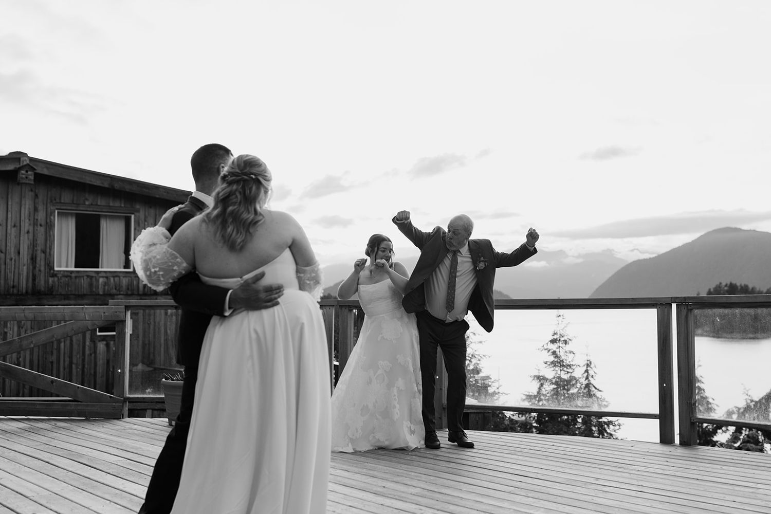 Wedding couples share a romantic moment on a scenic deck overlooking mountains and water at sunset.