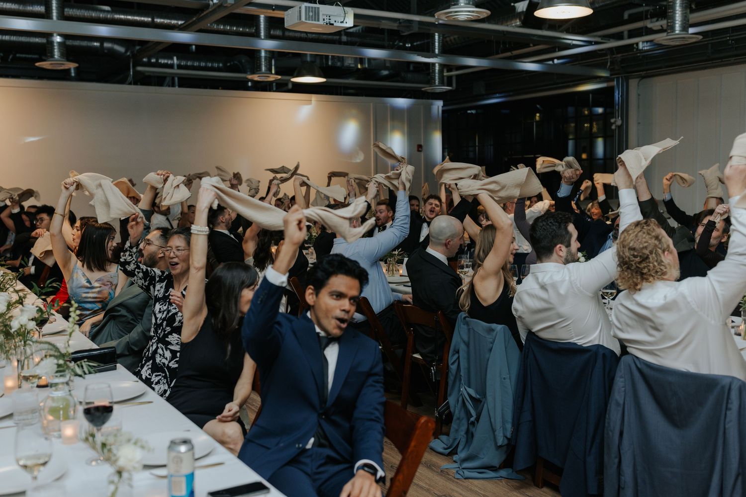 Wedding reception guests dancing and celebrating with arms raised in indoor venue.