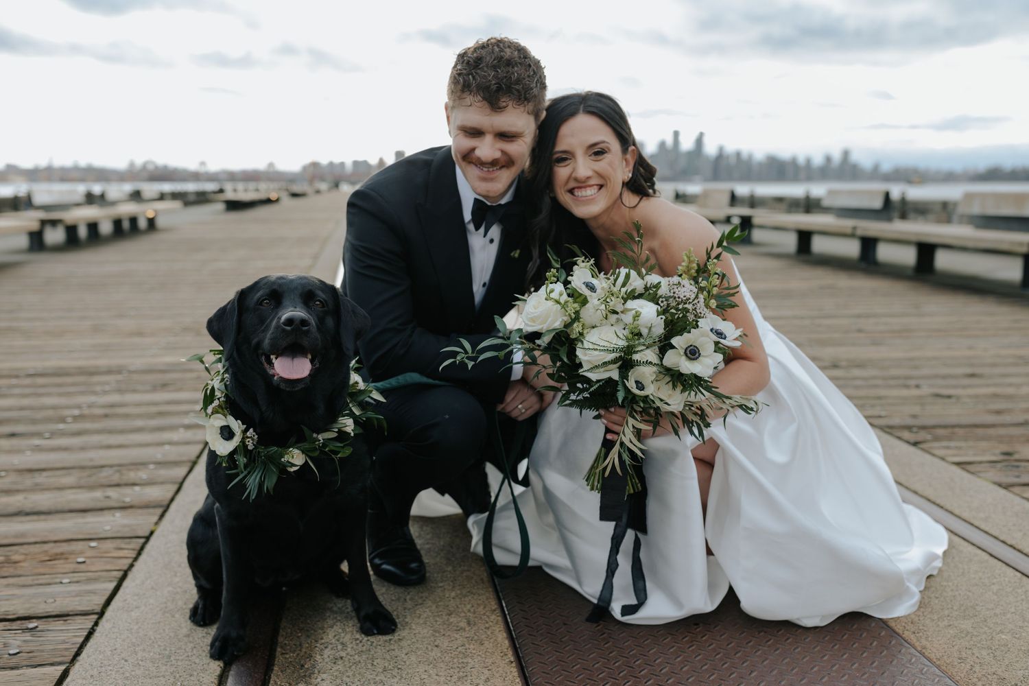 Wedding party poses with a black dog on a wooden pier with a city skyline in the background.