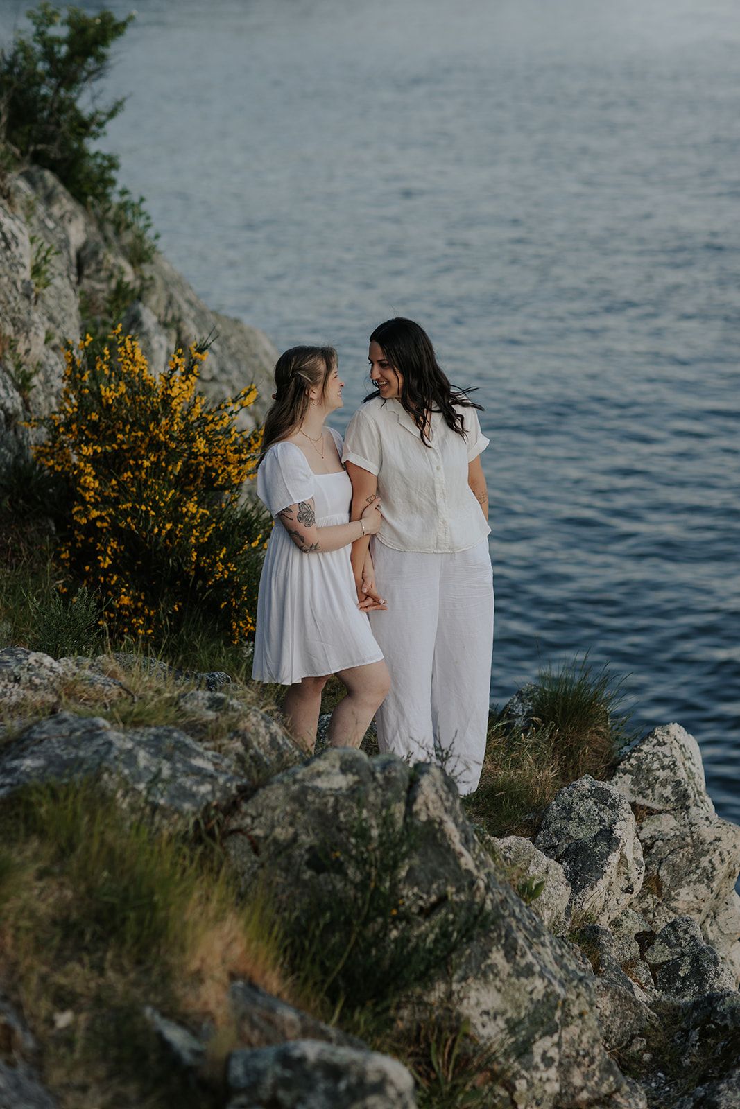 Two people in white dresses stand together on rocky coastal cliffs overlooking blue water.