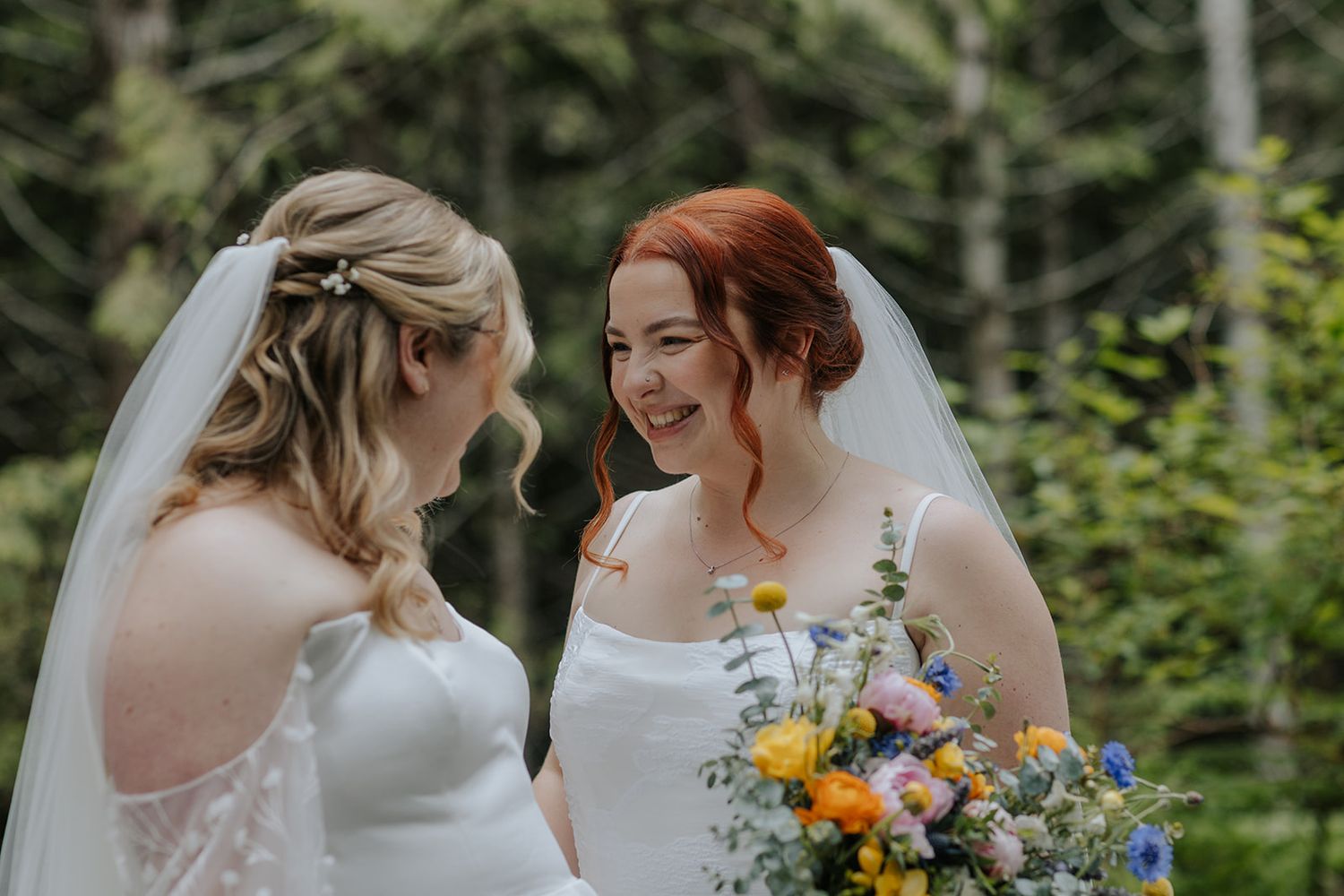 Two brides hold colorful wildflower bouquets while sharing happy moments in a forest setting.