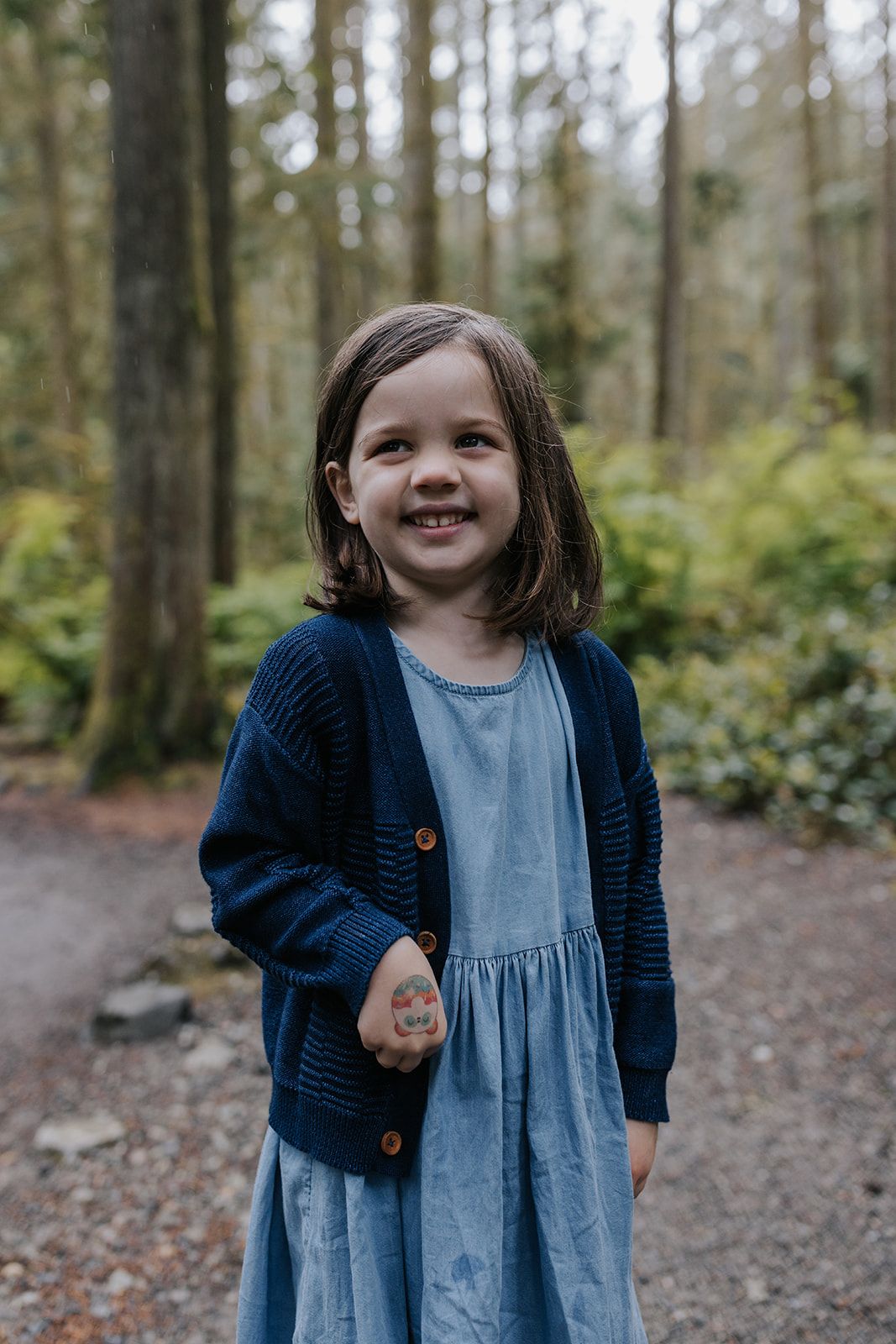 Young child in blue dress and cardigan smiling while playing in a forest setting.