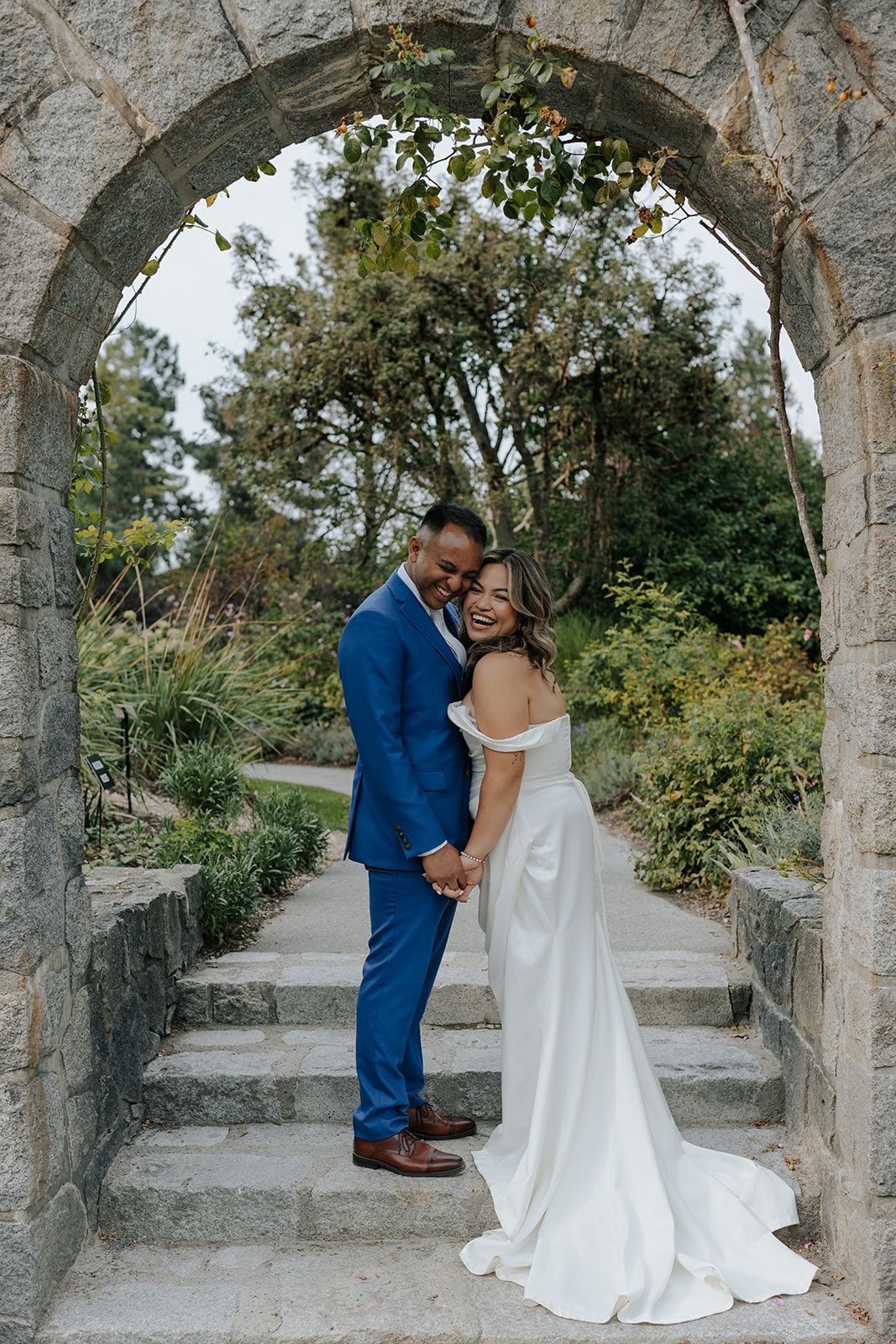 A couple shares romantic moments under a stone archway in a garden setting during their wedding celebration.
