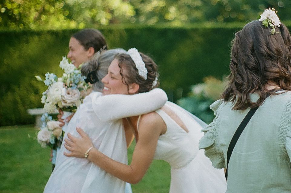 Wedding party members share an emotional embrace outdoors on a sunny day with white floral bouquets.