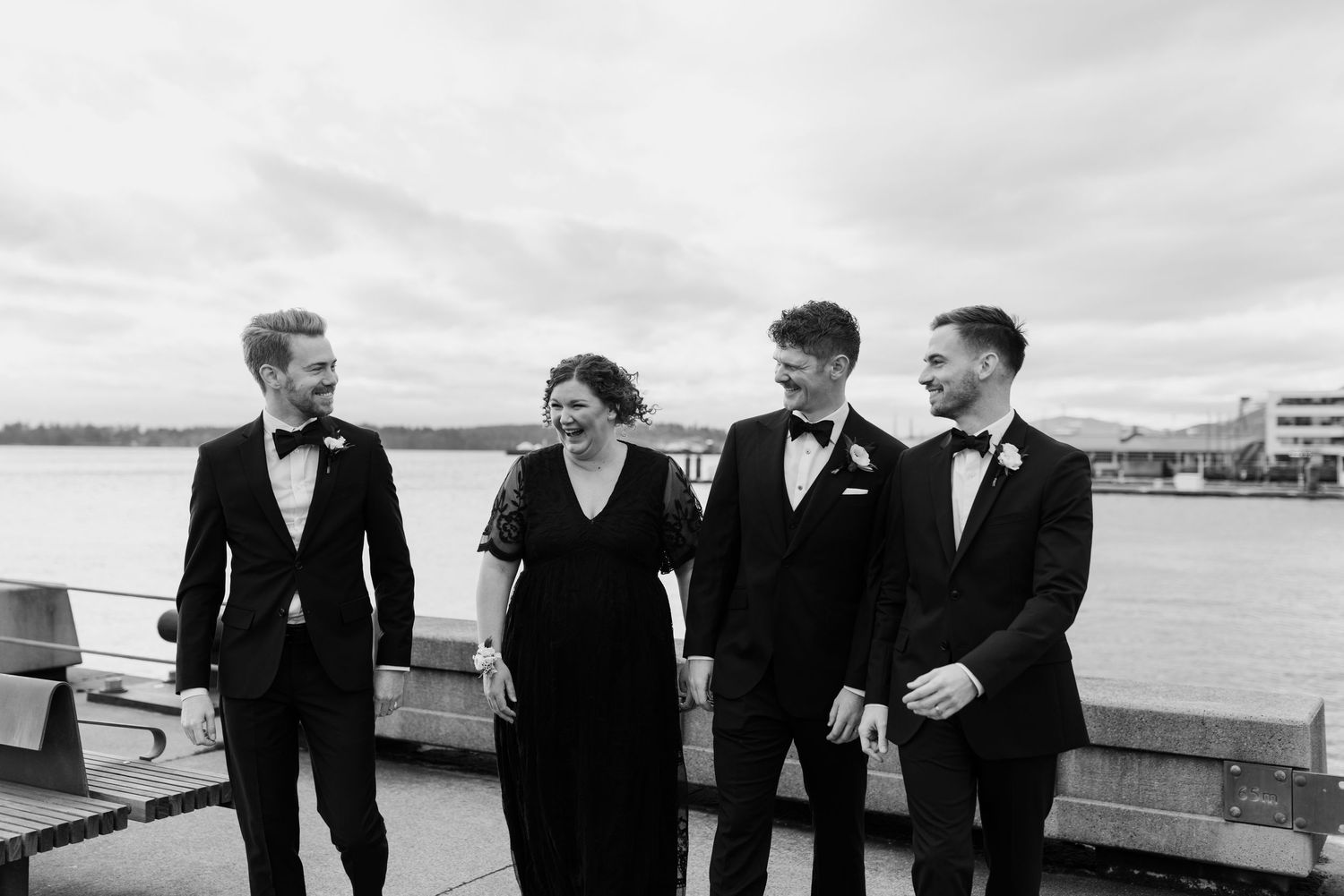 Wedding party in formal attire poses along waterfront railing with city skyline in background.