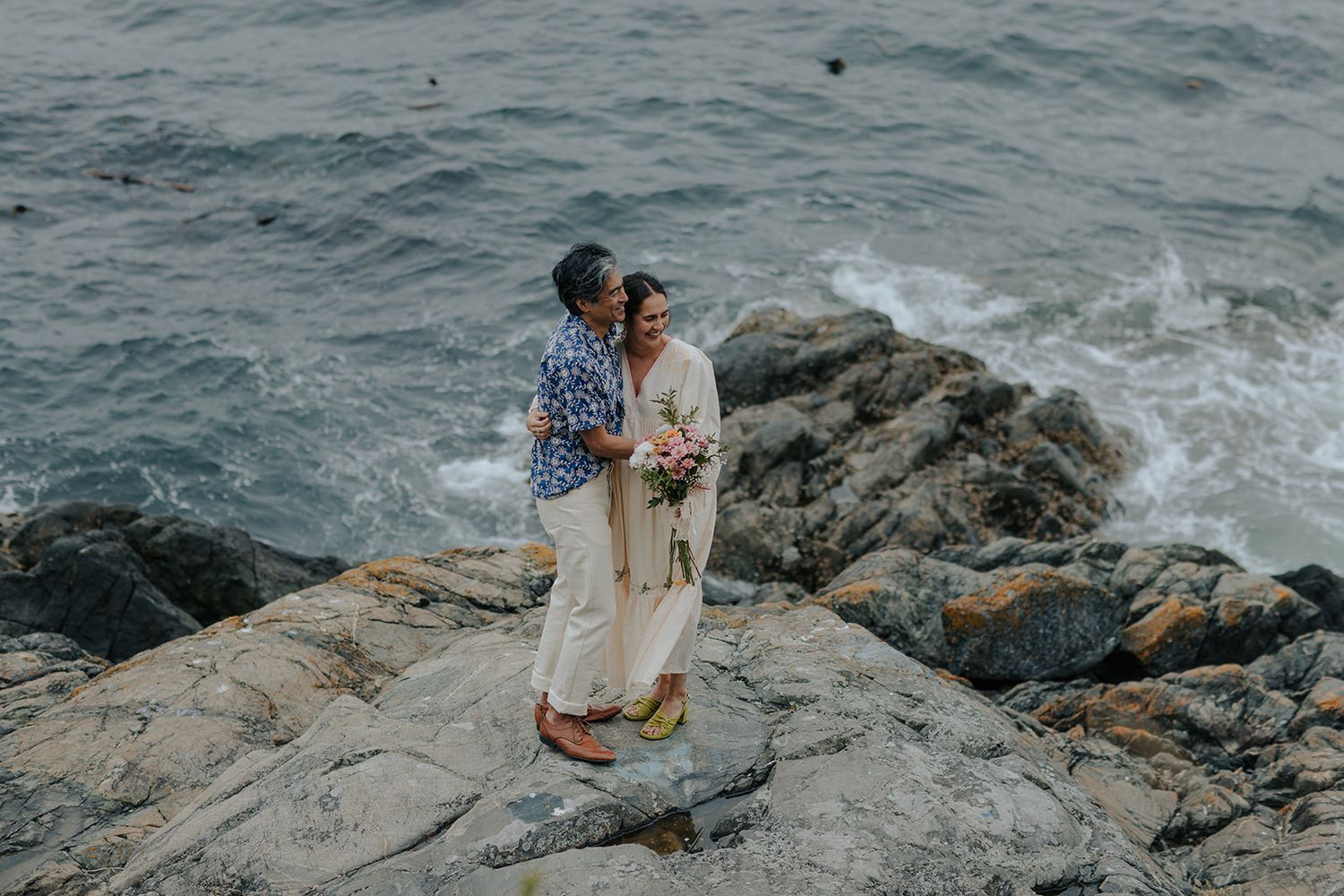 A couple embraces on rocky coastal cliffs overlooking the ocean during a cloudy day.