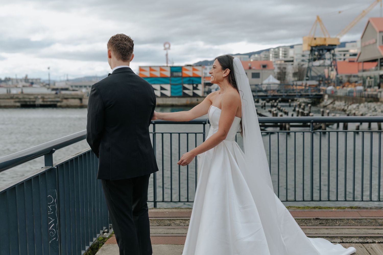 A couple in wedding attire standing on a pier overlooking a waterfront with boats and buildings visible.