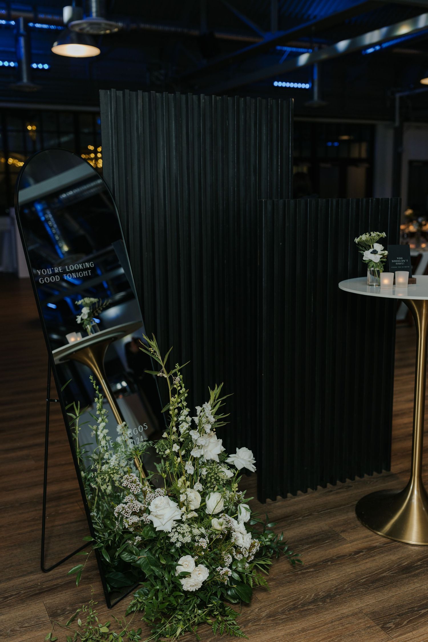 Elegant floral arrangements with white flowers and greenery decorate a dark venue with wooden flooring.