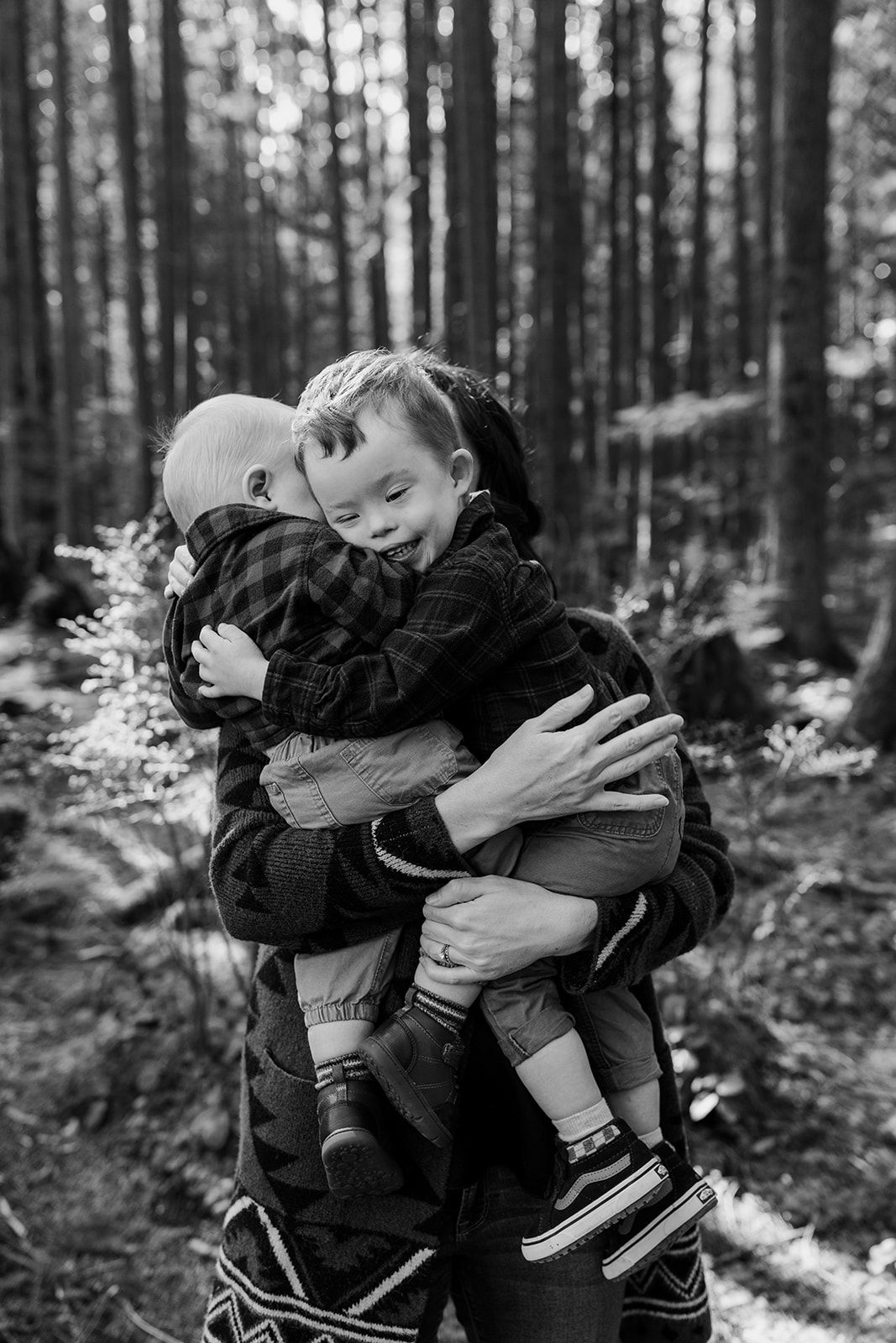 Black and white family portrait in pine forest showing joyful embrace.