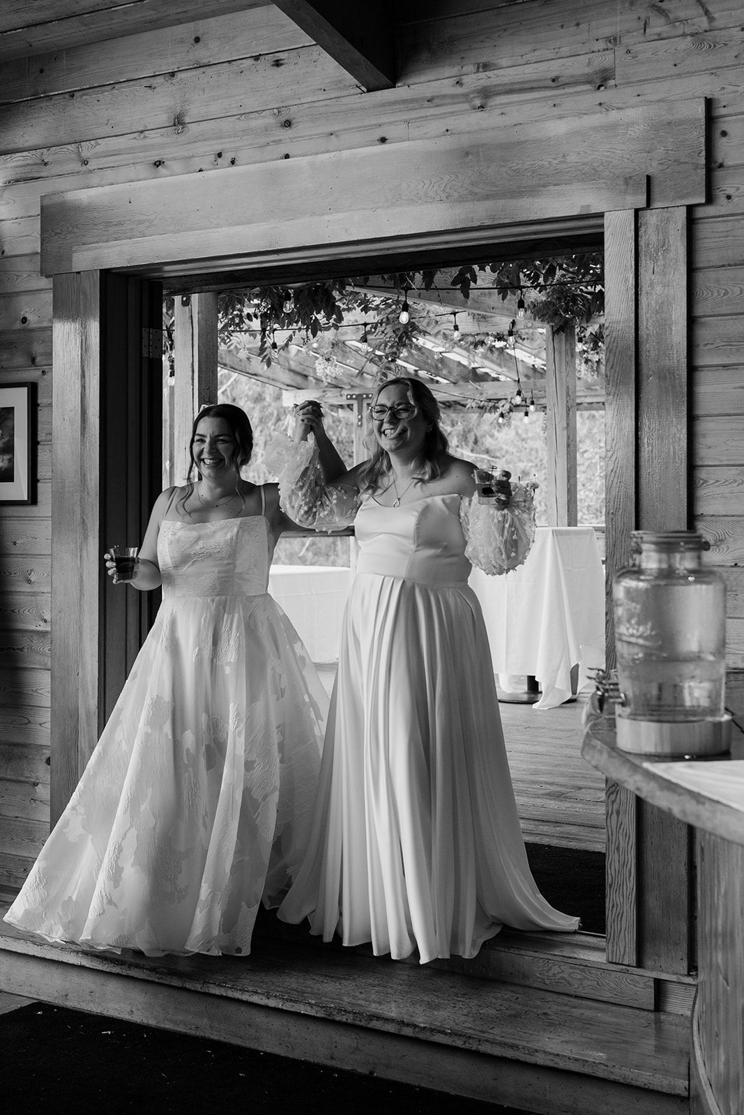 Black and white portrait of two brides in wedding gowns standing in a rustic doorway.