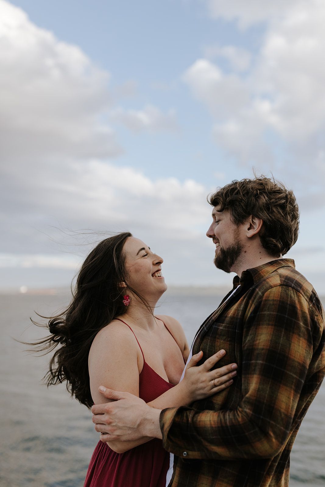 Couple embracing and laughing together against a dramatic cloudy ocean backdrop.
