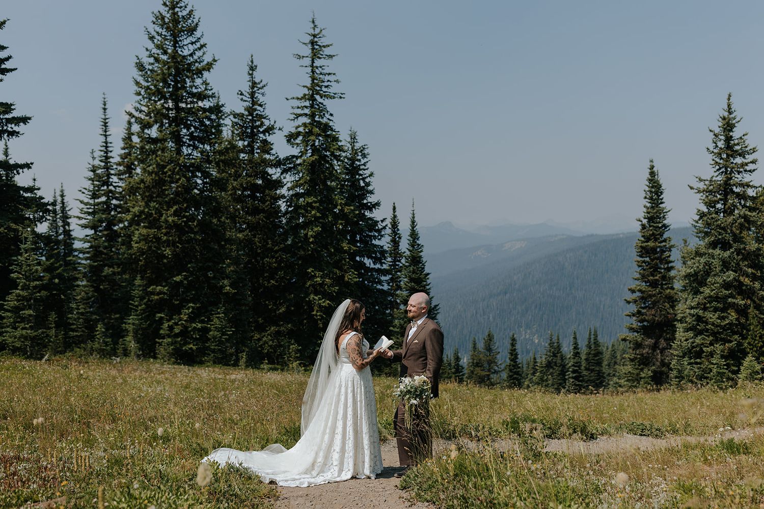 A wedding couple poses in a scenic mountain meadow surrounded by evergreen trees.