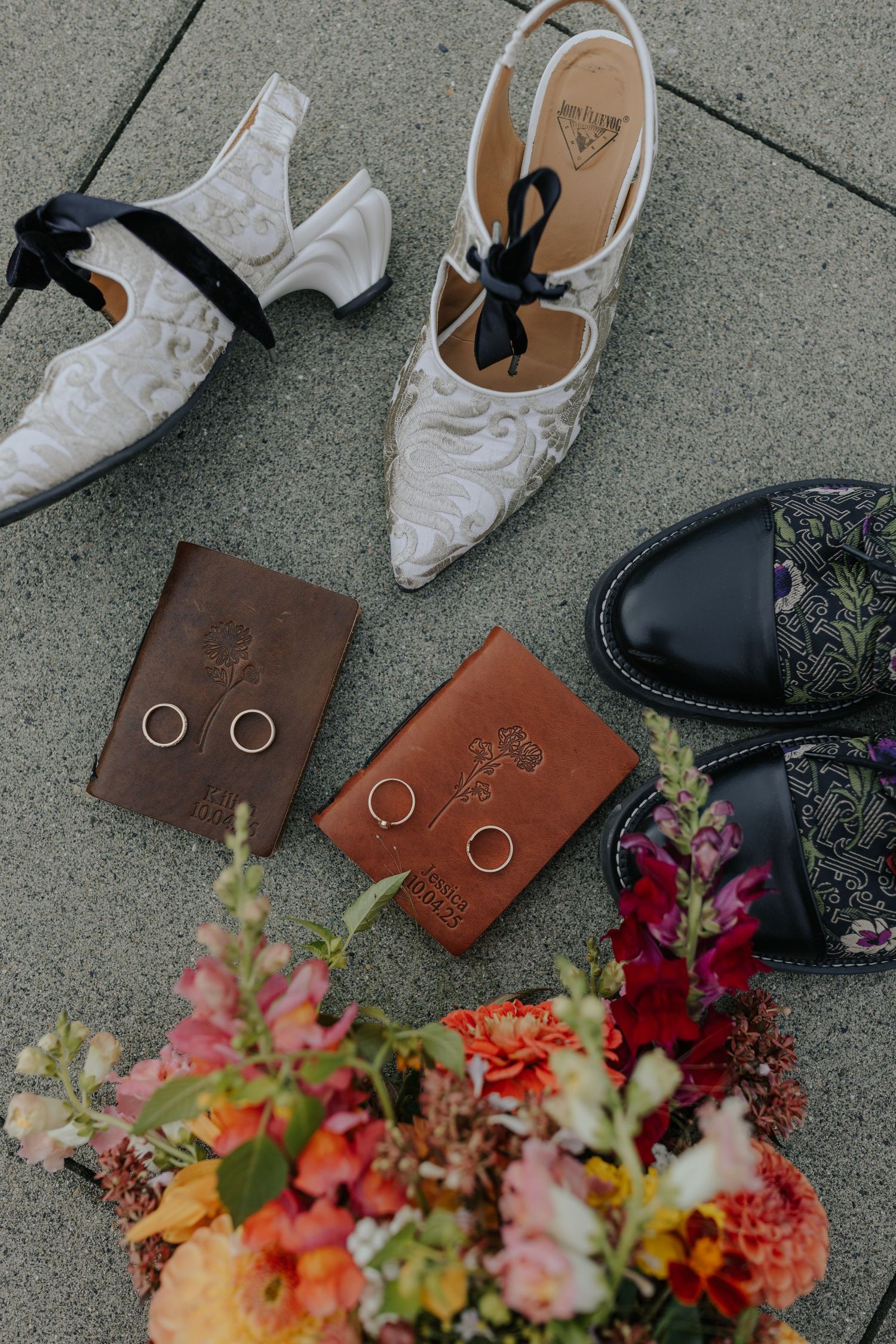 Wedding details showing rings on leather wallets with colorful floral bouquet and shoes on concrete.