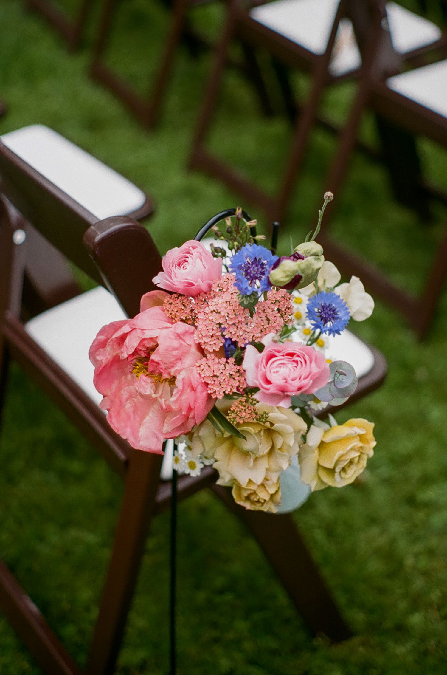 Colorful floral decorations featuring pink, blue, and cream flowers adorn wooden chairs at an outdoor event setting.